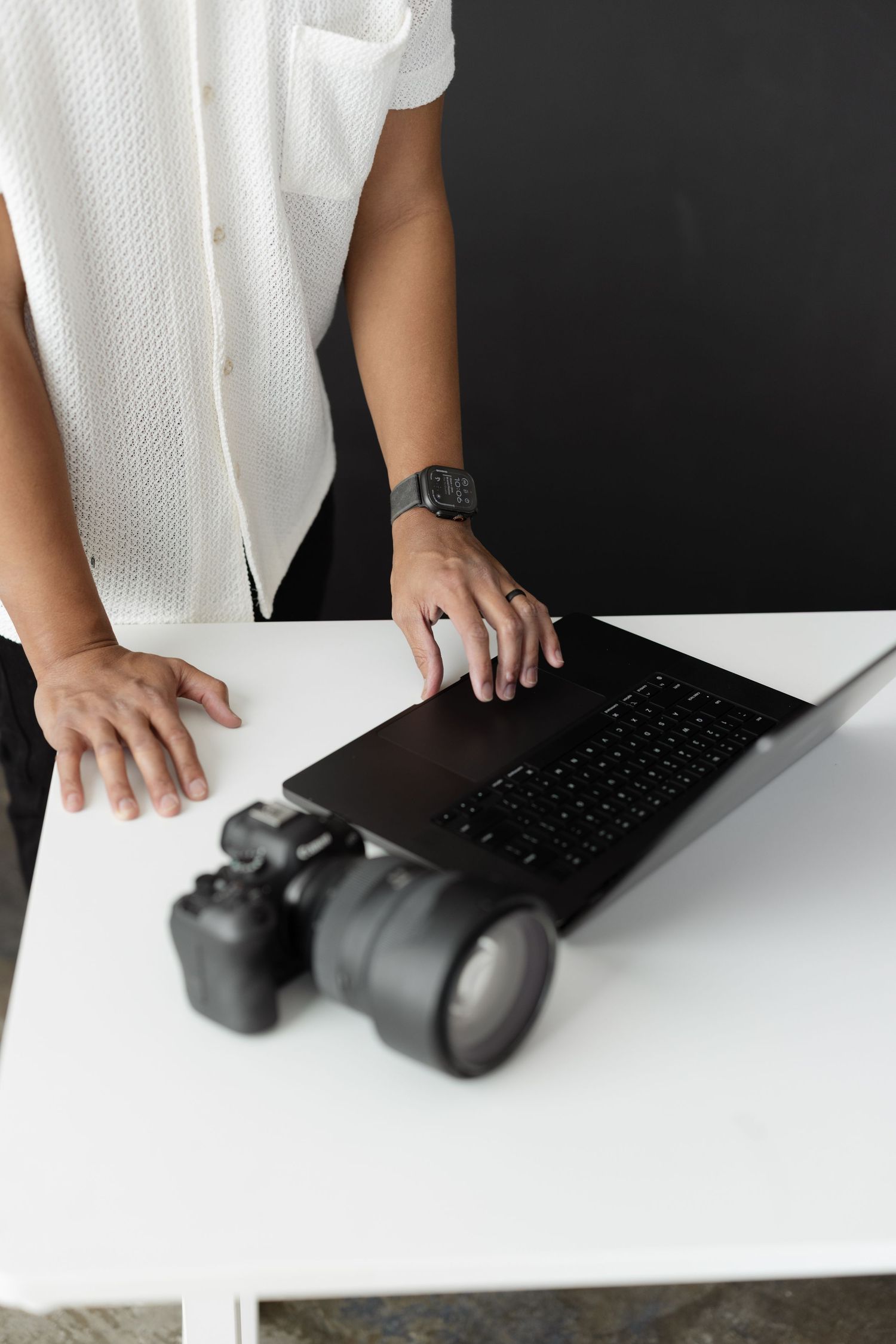Photographer working on laptop beside DSLR camera on white desk against dark background.