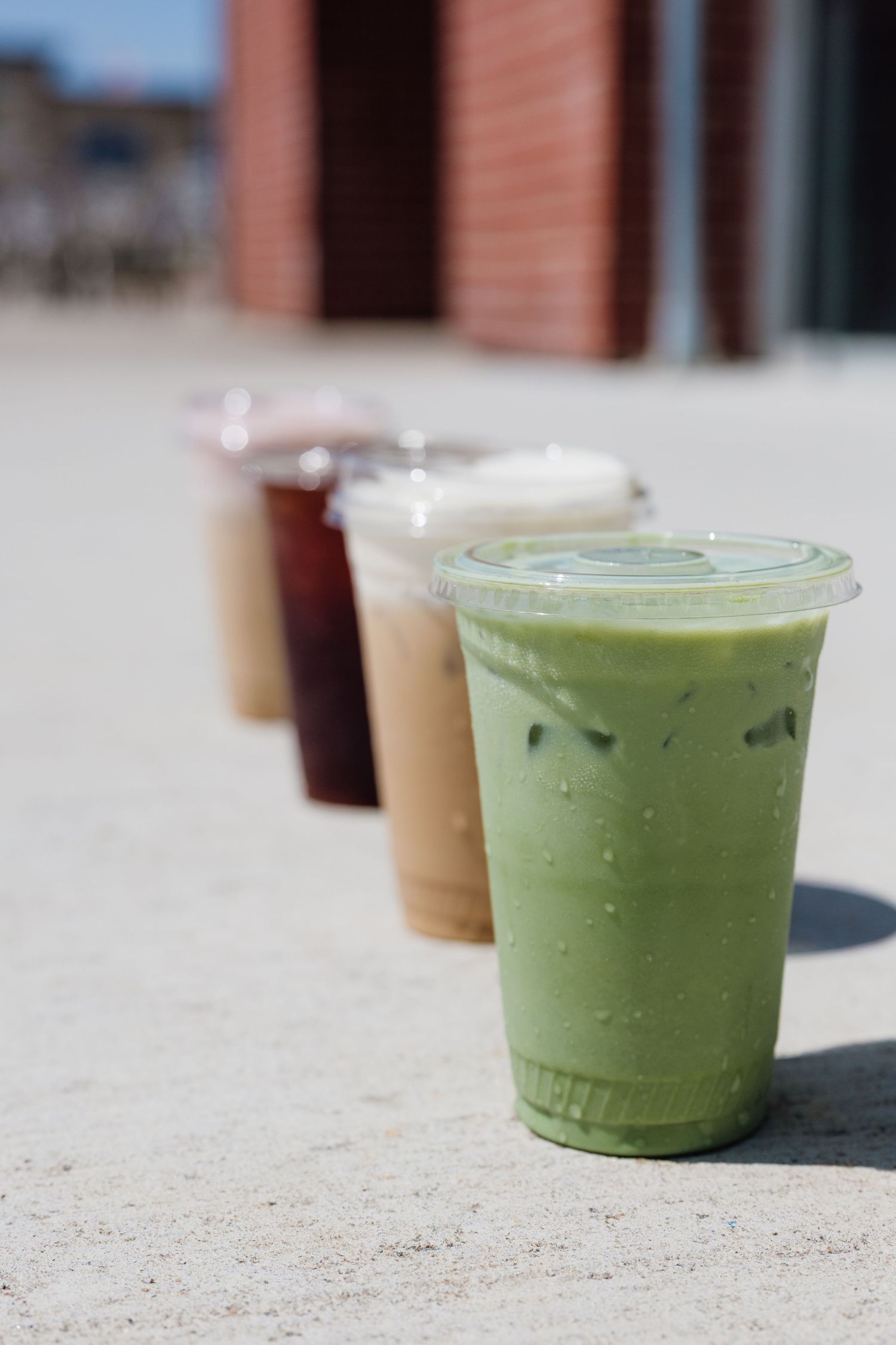 Row of iced drinks in plastic cups outdoors, featuring matcha latte, coffee, and dark beverages on a sunny day.
