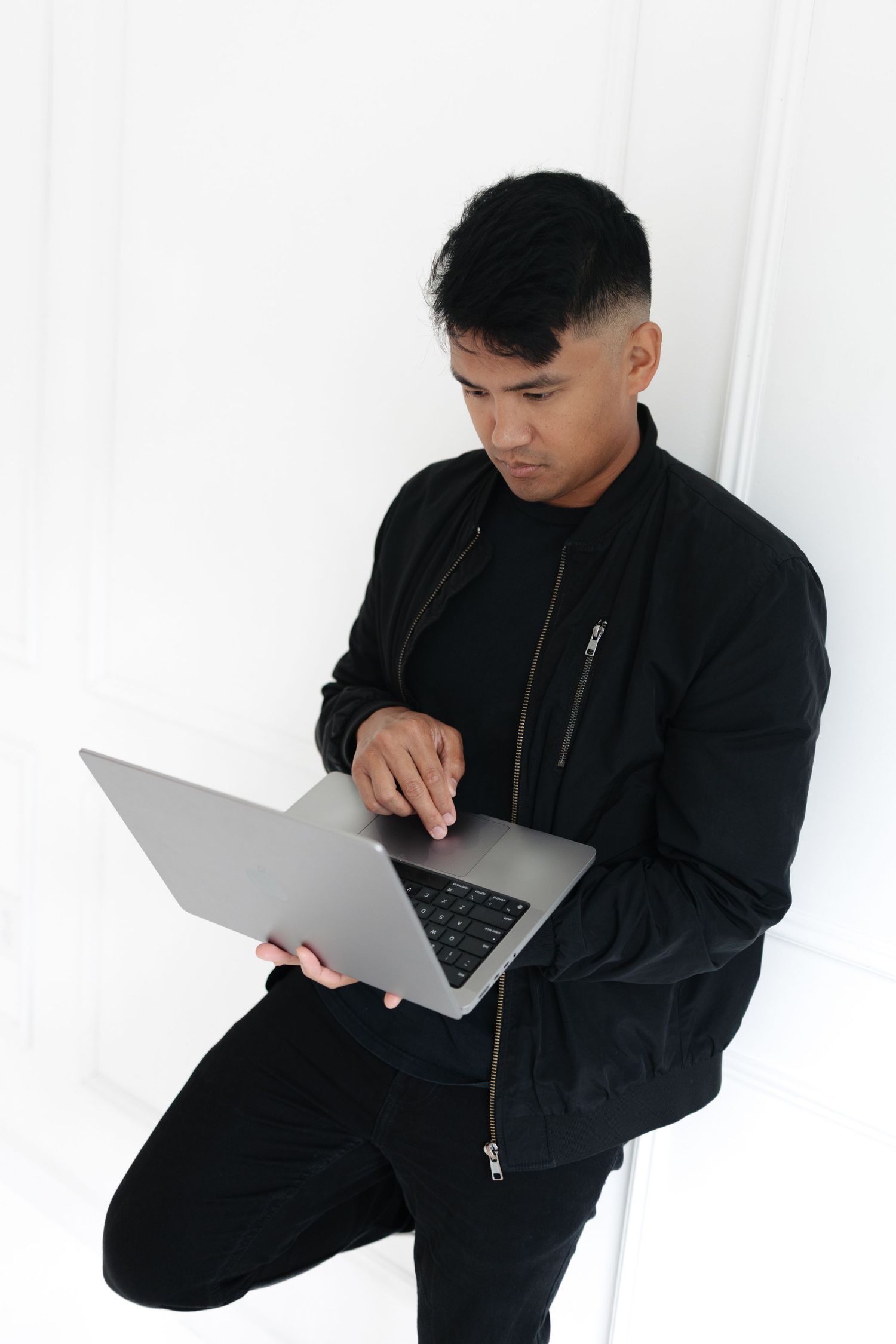 Young man in black jacket using laptop computer while standing against white wall.