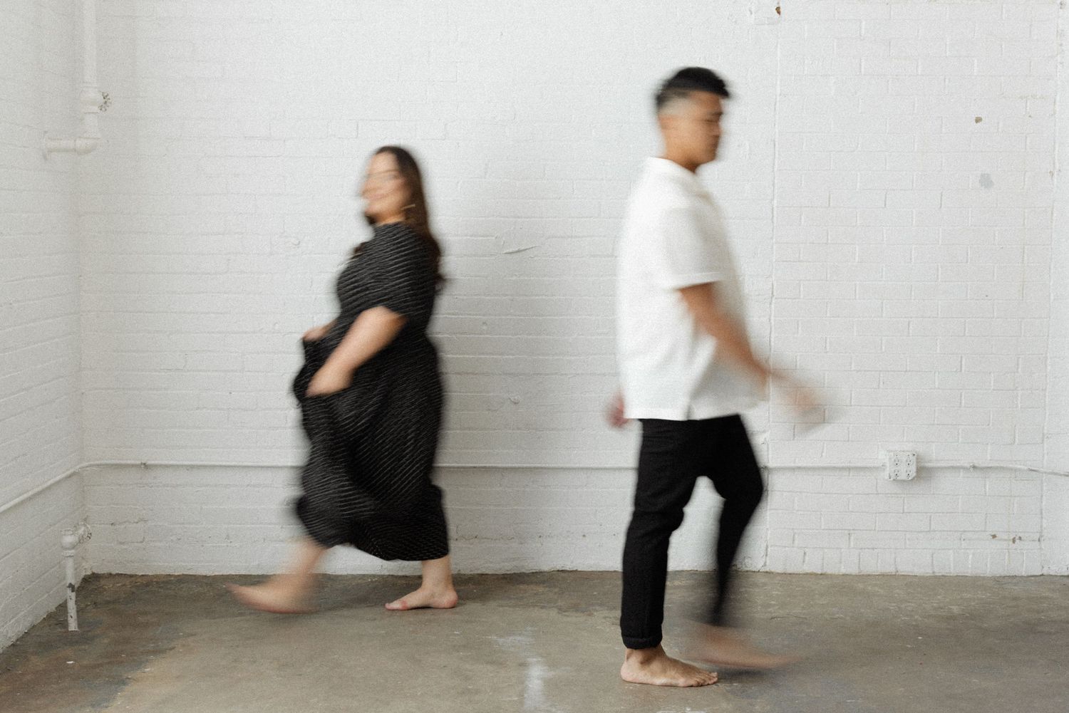 A man and woman walk barefoot in a white brick studio, captured with motion blur in black and white outfits.