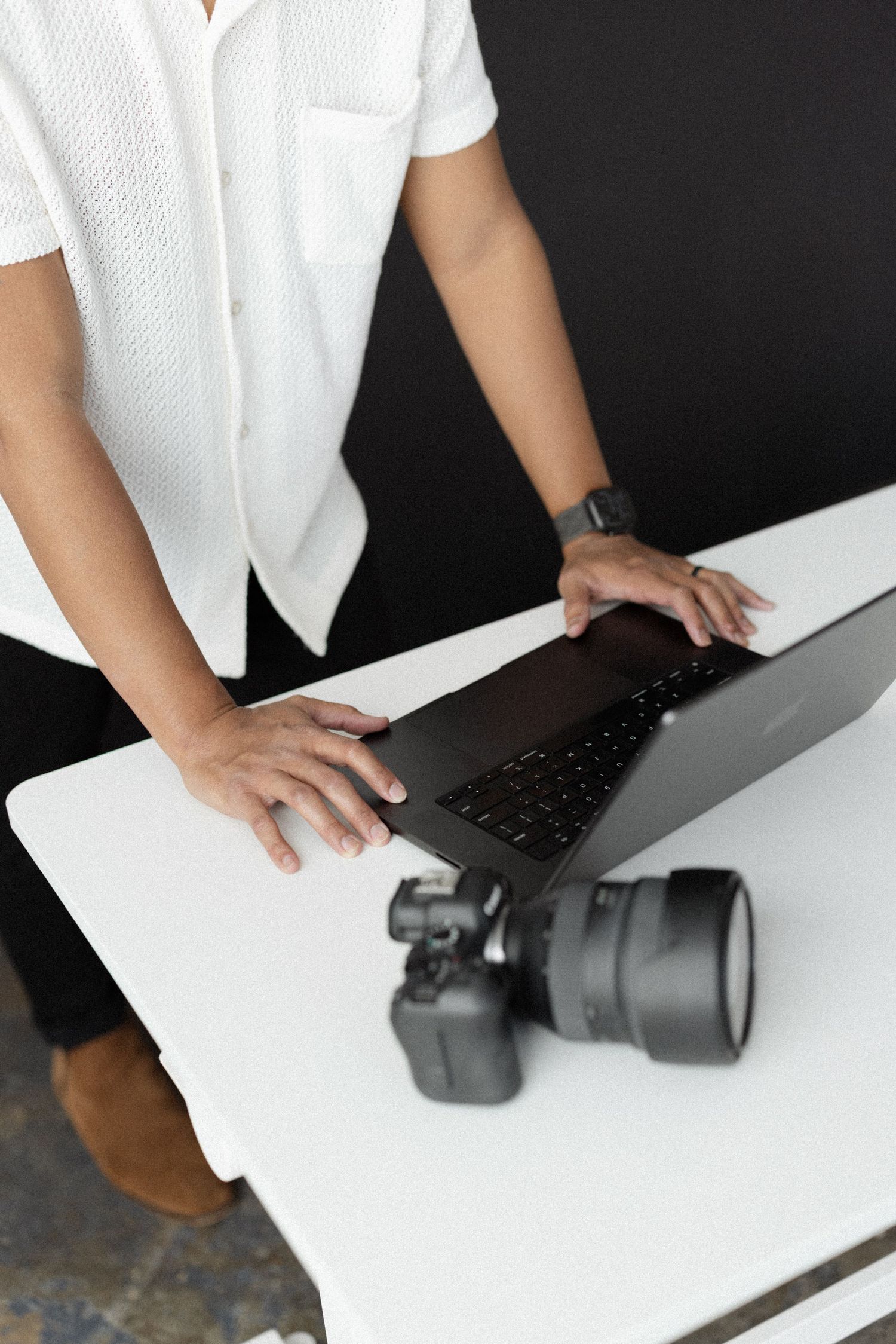 Overhead view of person in white shirt using laptop next to DSLR camera on white table.