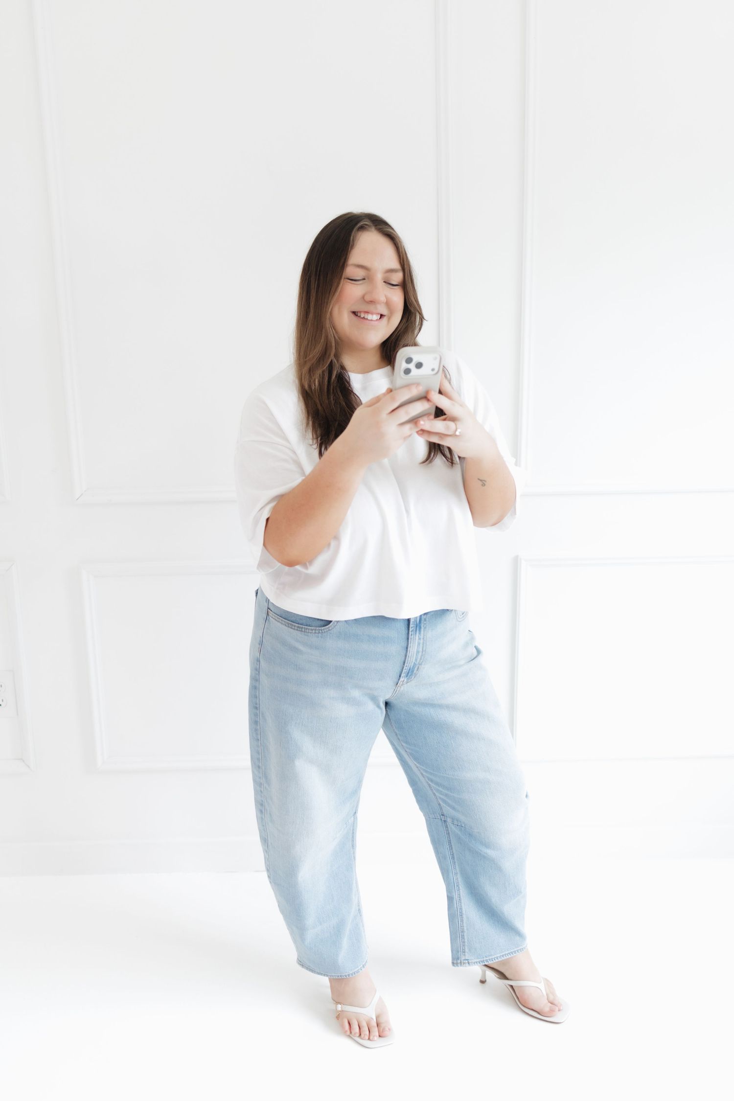 A smiling plus-size woman in a white crop top and light blue jeans holds a smartphone in a bright white studio.