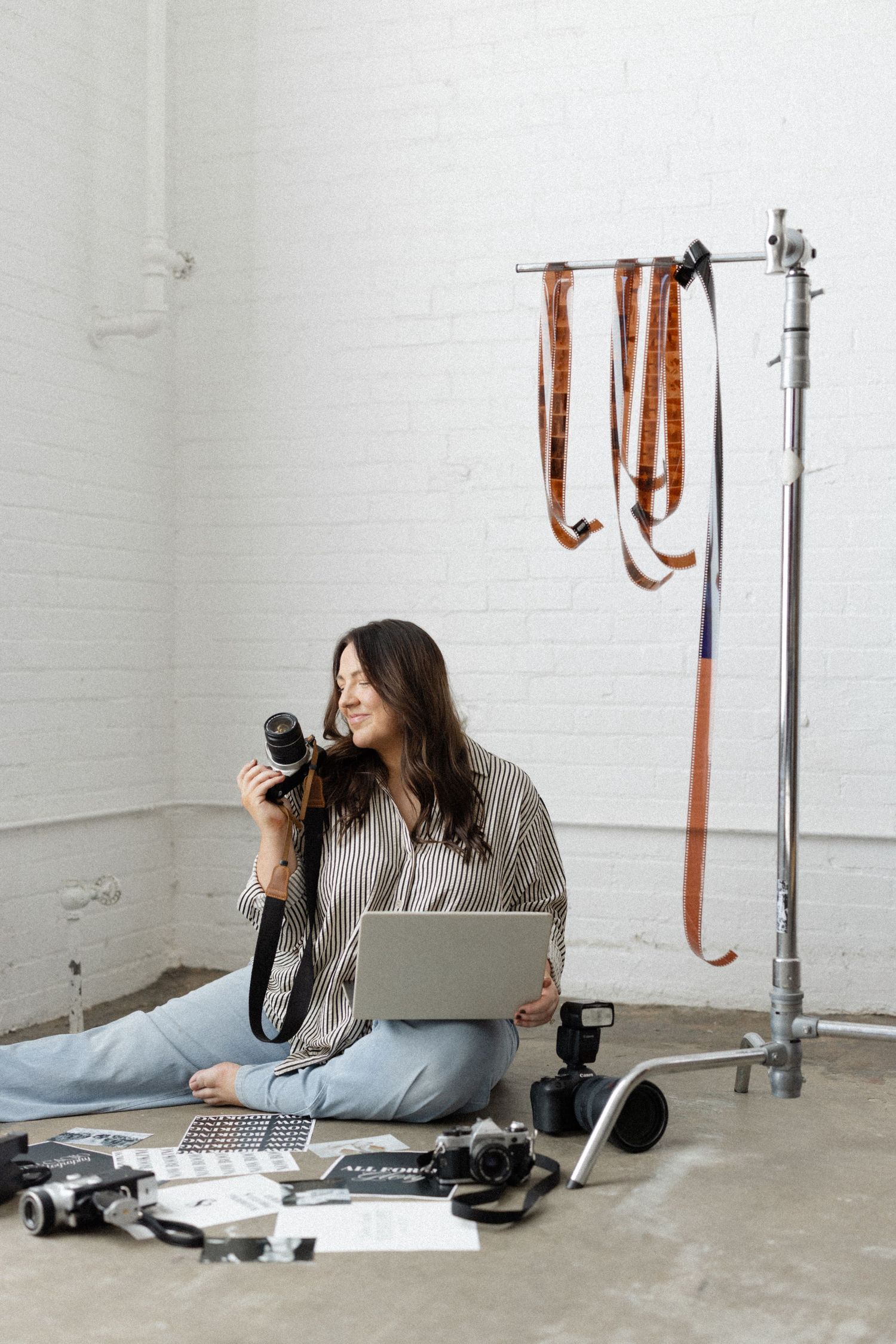 Female photographer sitting on studio floor with camera, laptop, and film strips in creative workspace.