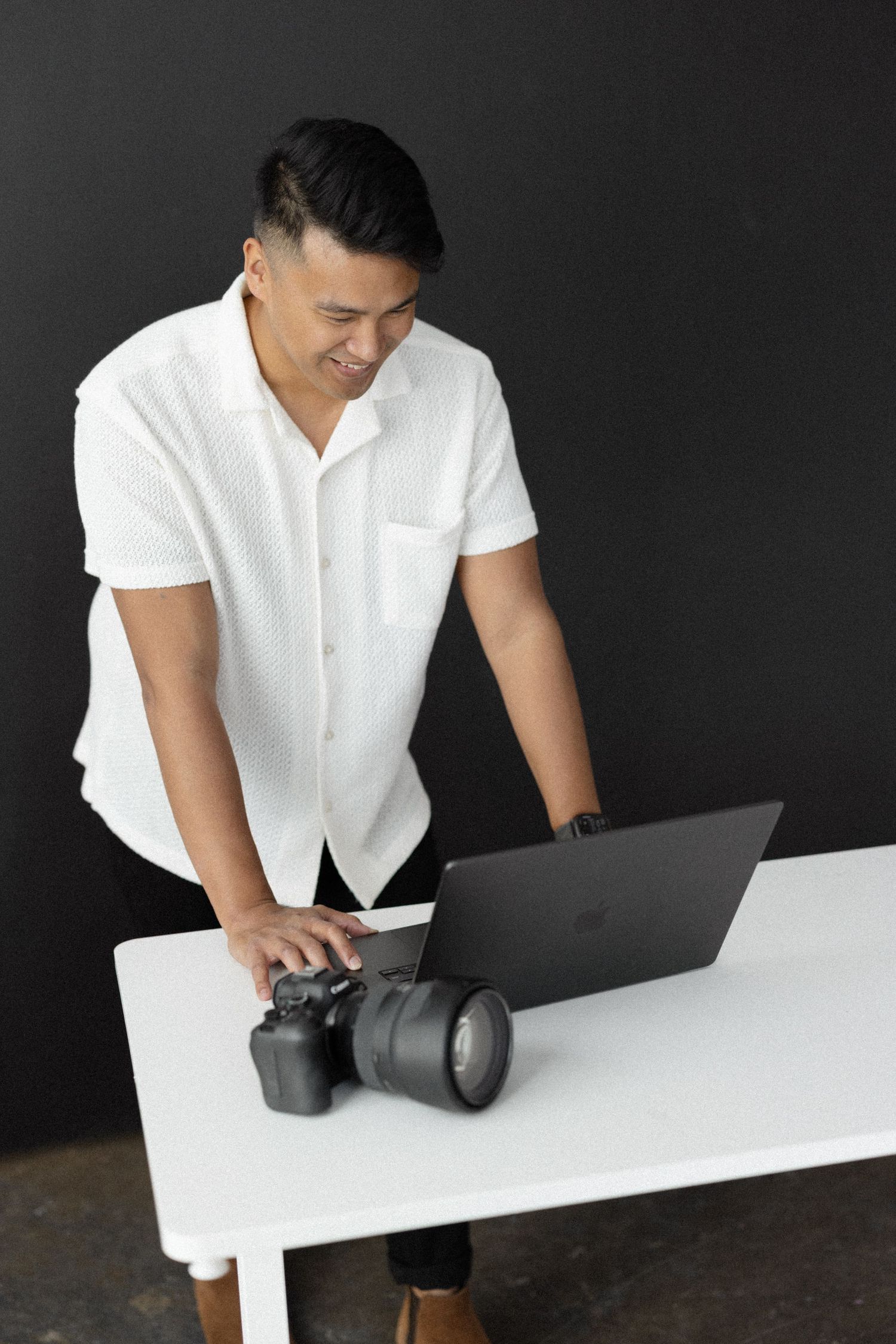 Man in white shirt smiling while working on laptop with DSLR camera on white table against dark background.