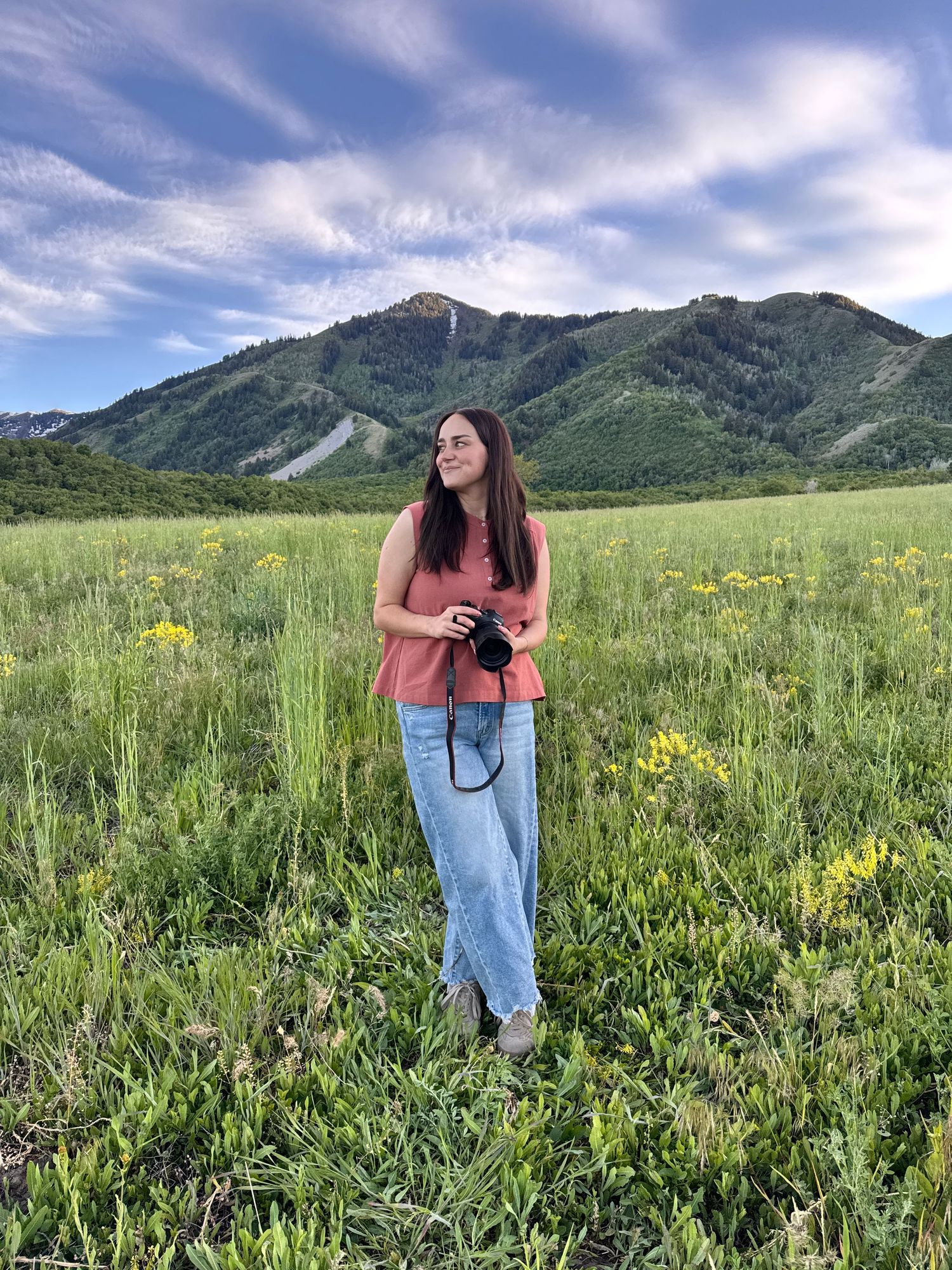 Woman holding camera stands in green meadow with yellow wildflowers, mountains and blue sky behind her.