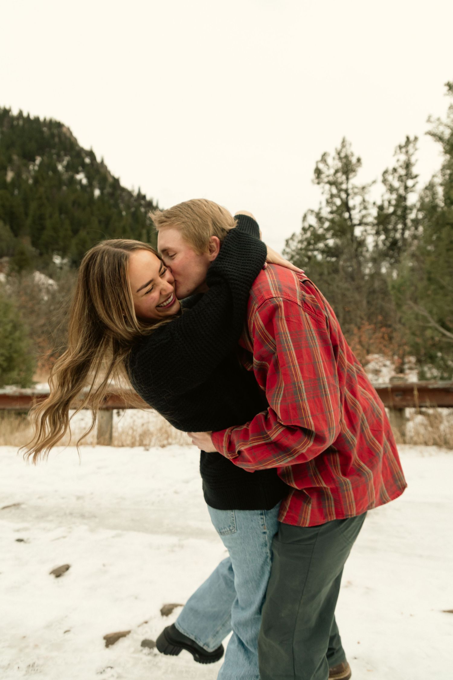 Couple embracing and laughing in snowy winter landscape with pine trees, one wearing red plaid shirt.