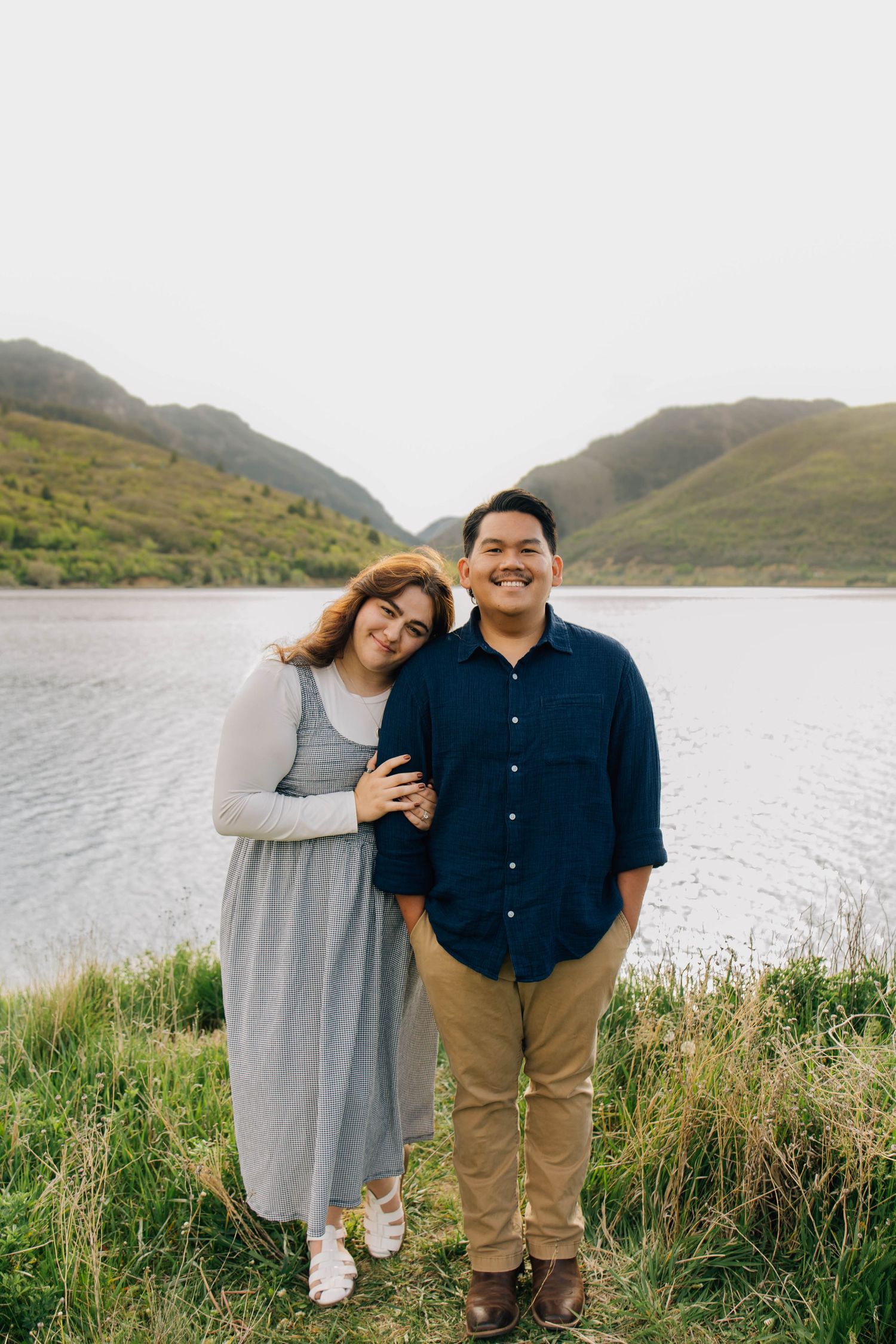 Couple standing close together by a mountain lake, woman resting head on mans shoulder, smiling warmly.