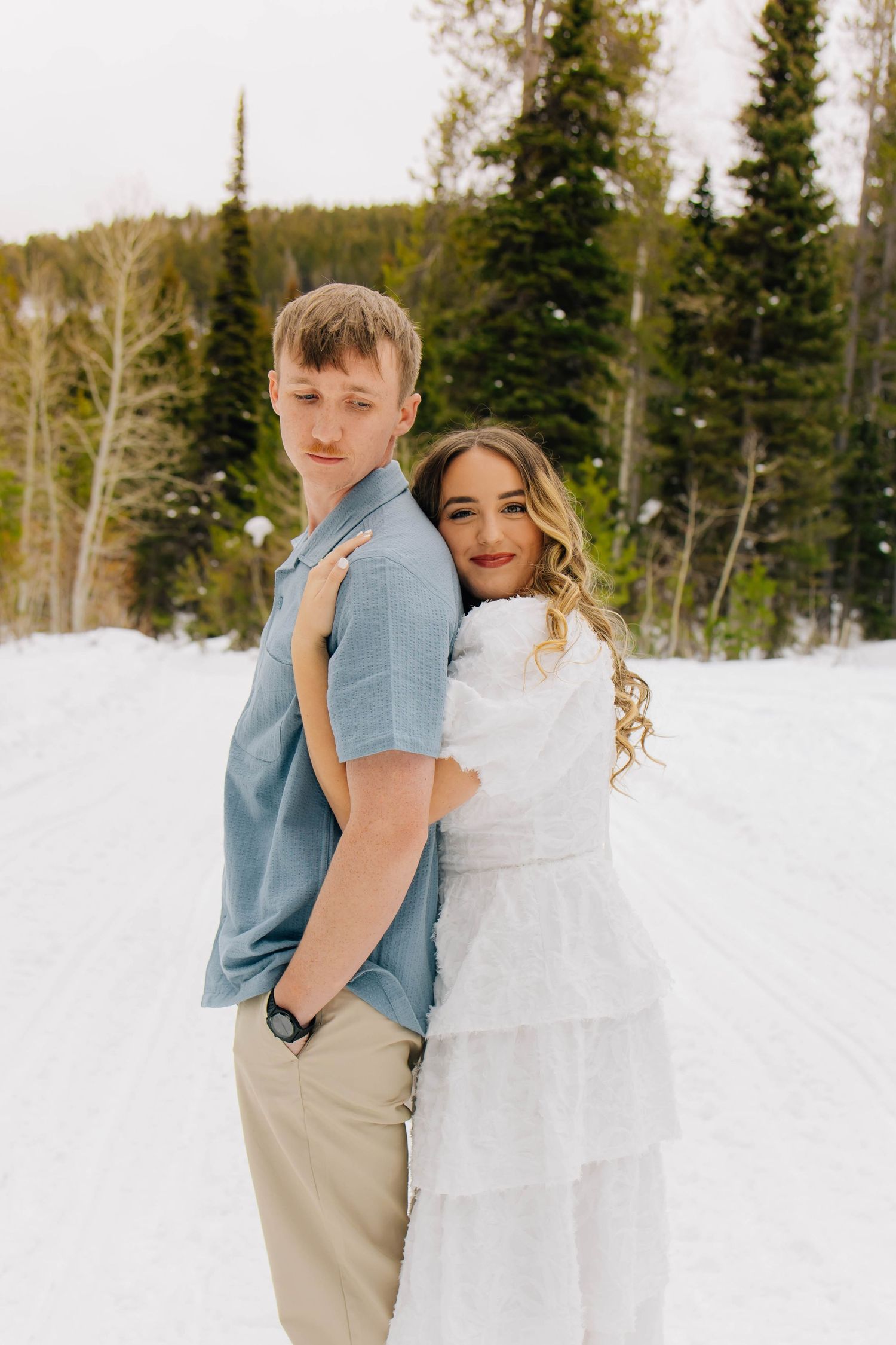 Couple posing in snowy winter forest, woman in white dress hugging man in blue shirt from behind.