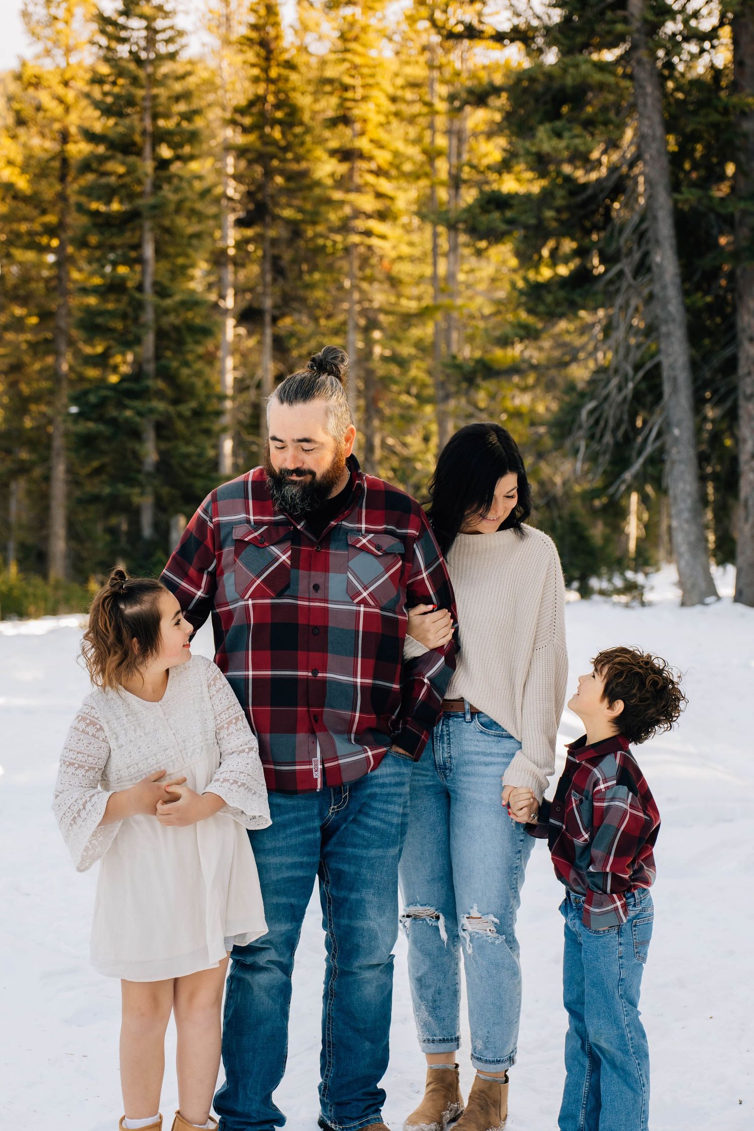 Family of four in coordinating plaid outfits walking together in snowy pine forest during winter.
