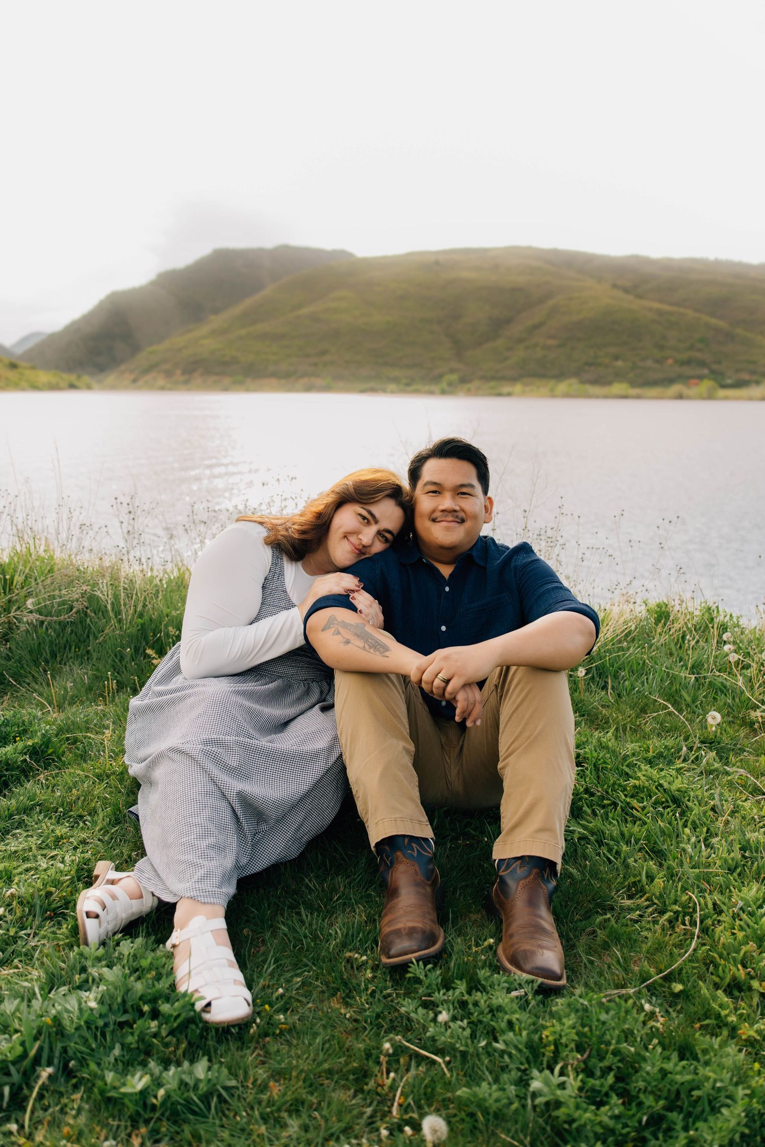 Couple sitting together on grass by a misty lake with green hills, woman leaning on mans shoulder smiling.