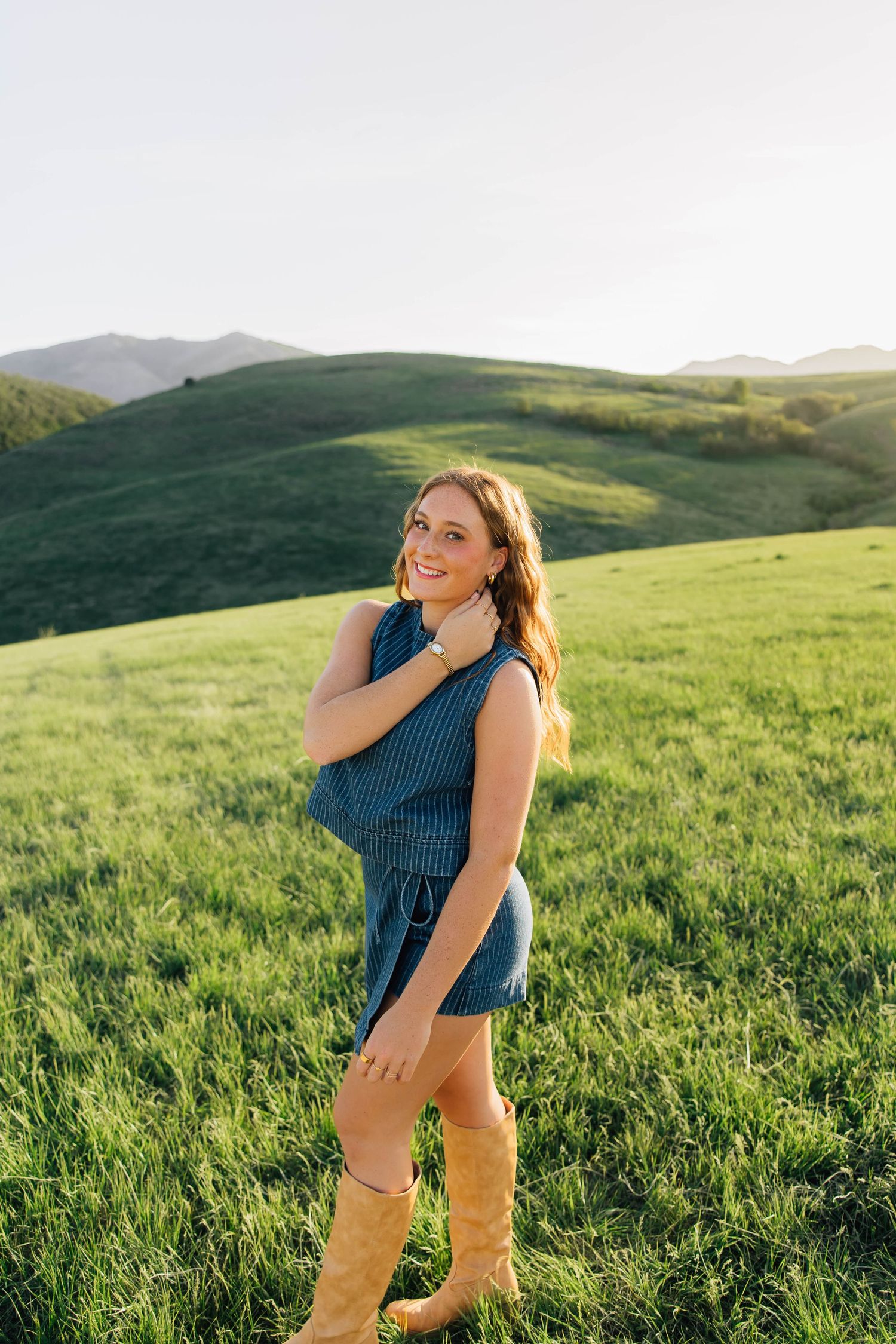 Young woman in teal dress and cowboy boots smiling in a lush green field with rolling hills backdrop.