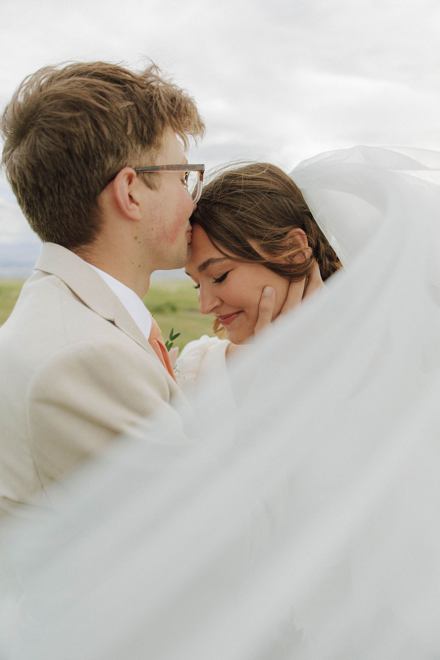 Groom kissing brides forehead as flowing white veil wraps around them in romantic outdoor wedding portrait.