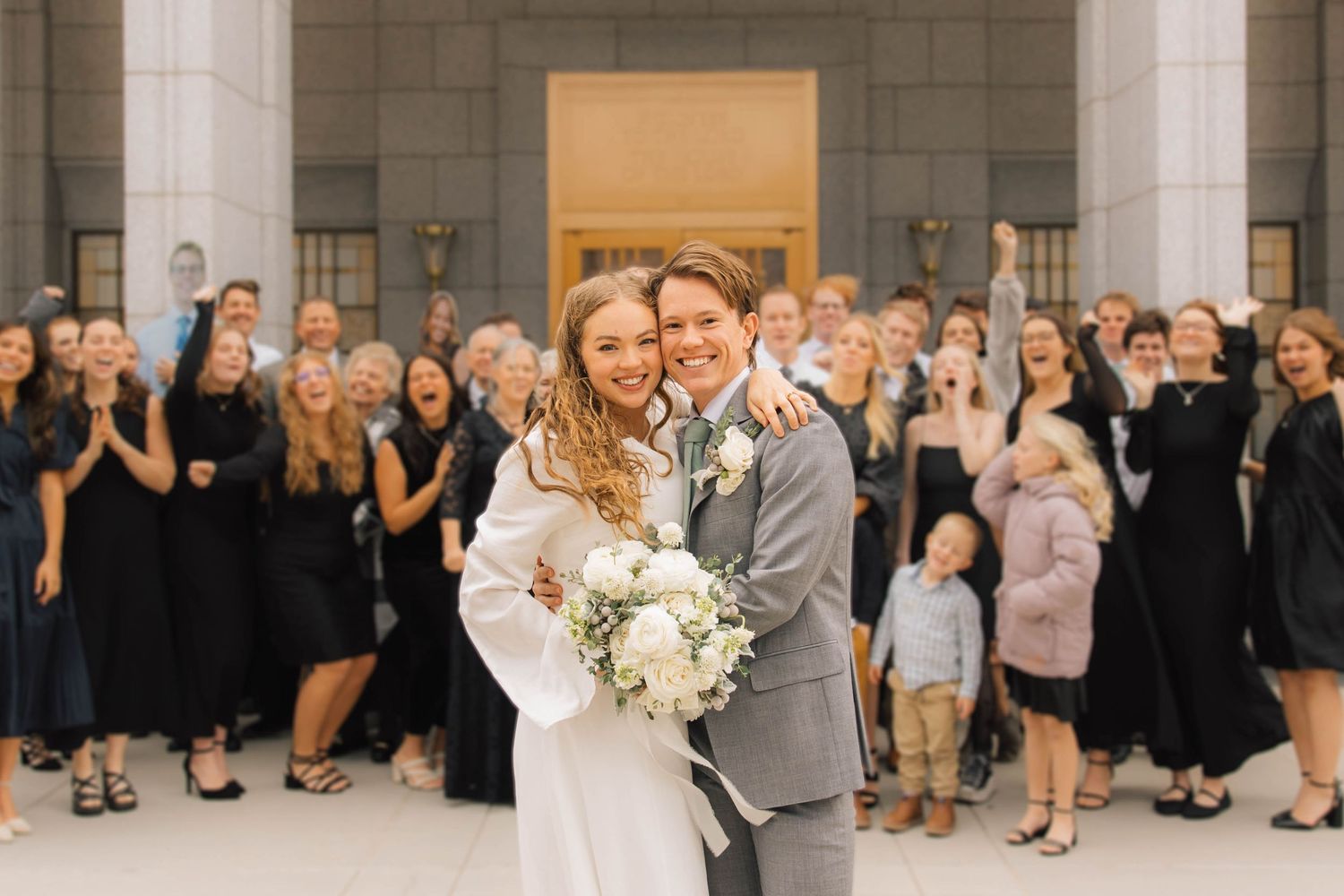 Bride and groom hugging joyfully outside temple steps surrounded by cheering wedding guests.