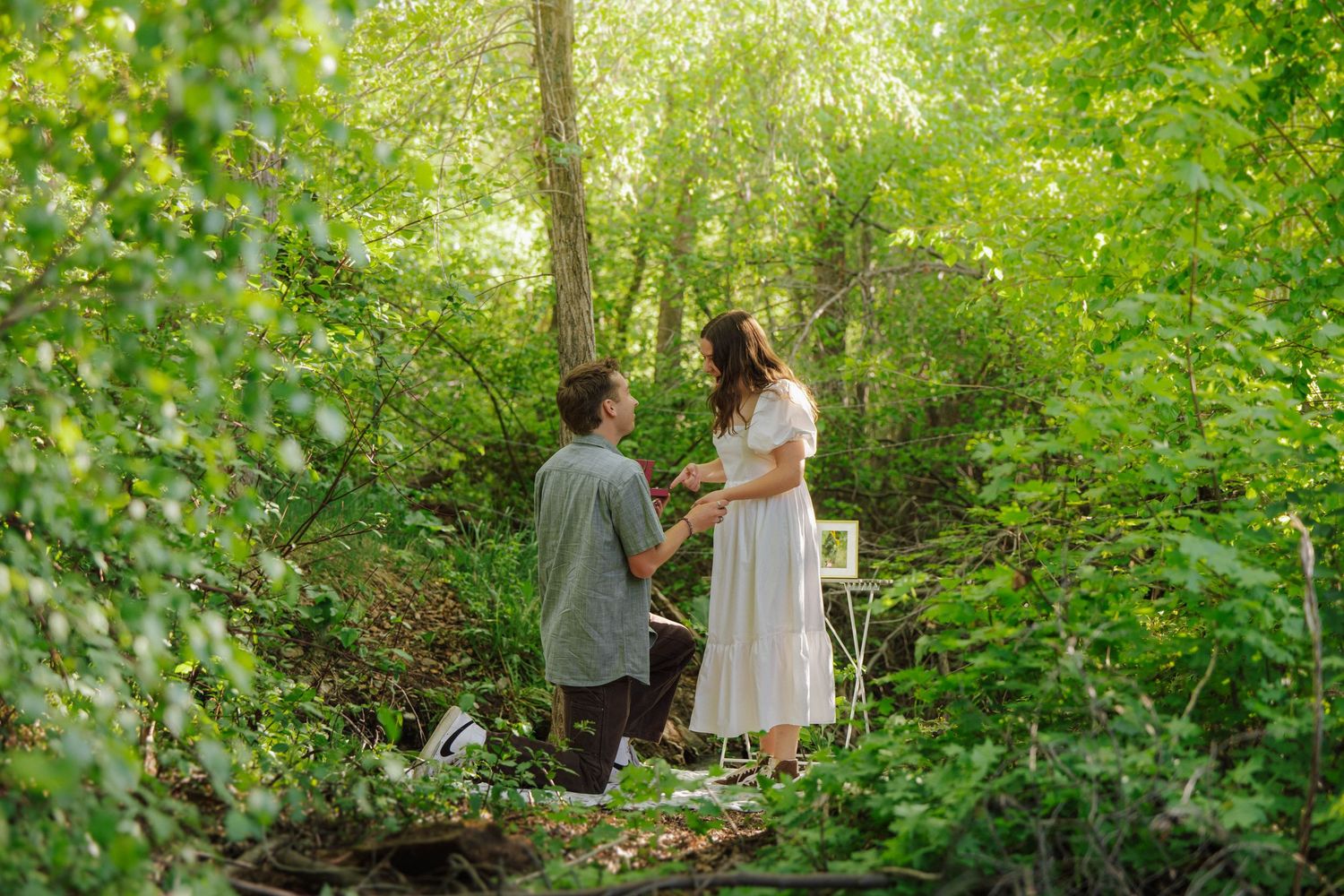 Man kneeling in proposal to woman in white dress in lush green forest with decorative table nearby.