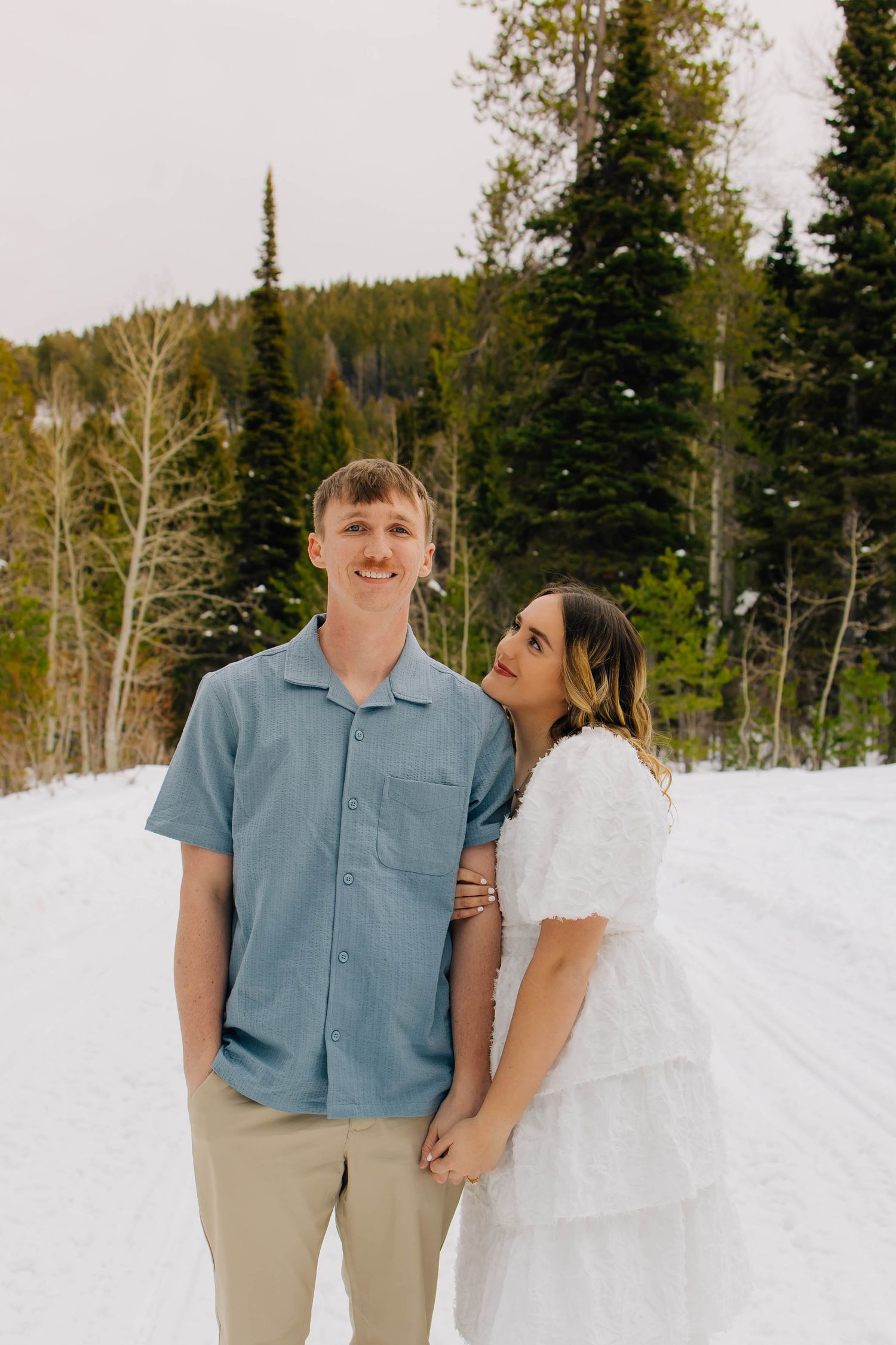 Smiling couple holds hands on a snowy mountain road surrounded by pine and aspen trees.