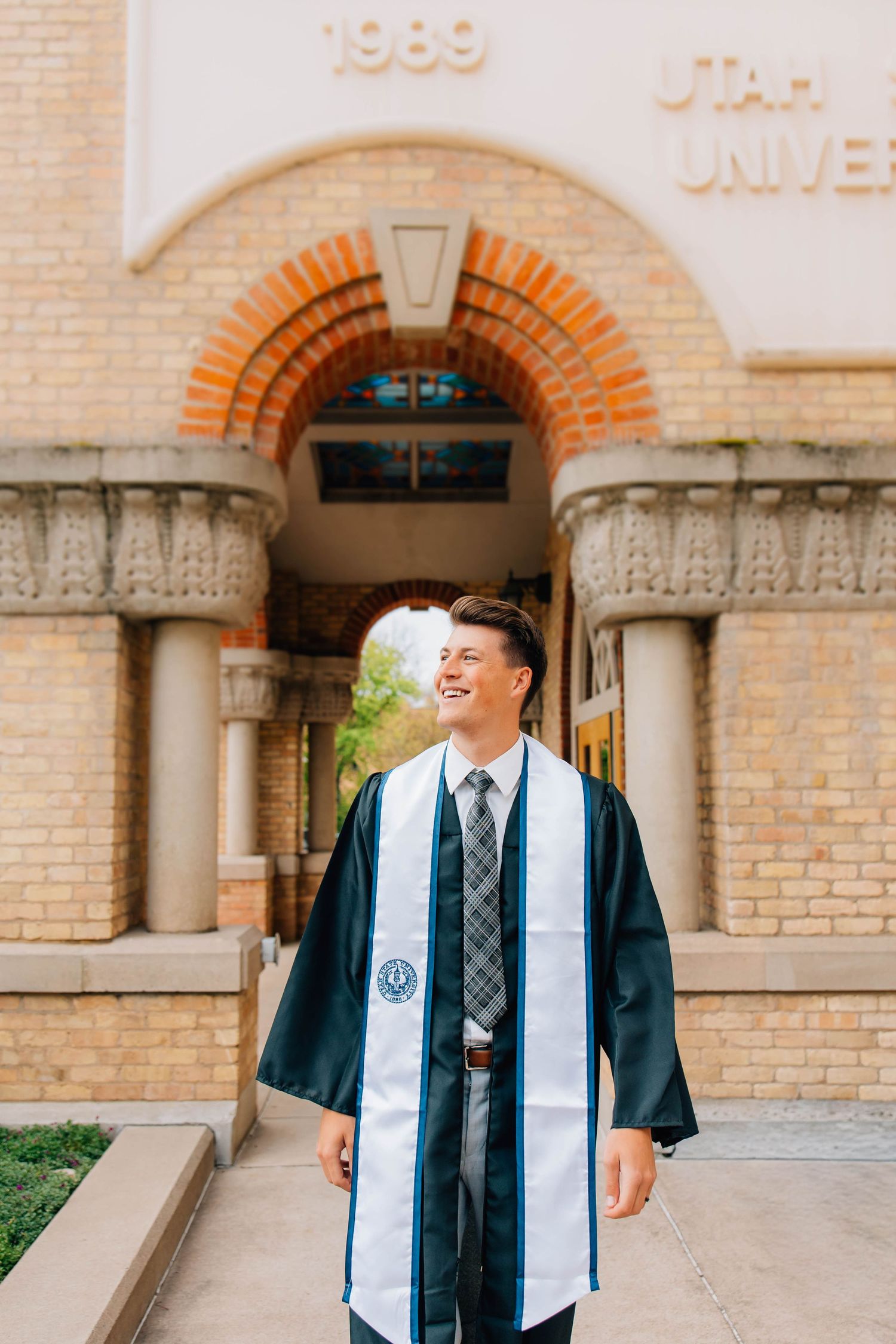 Smiling male graduate in cap and gown stands before Utah State University brick archway.