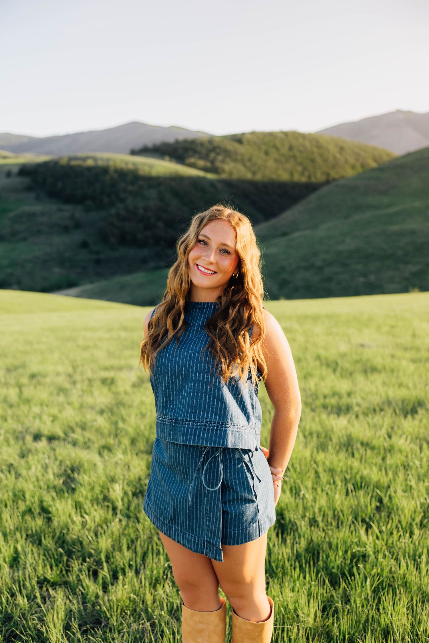 Smiling young woman in blue striped romper and tan boots posing in green rolling hills at sunset.