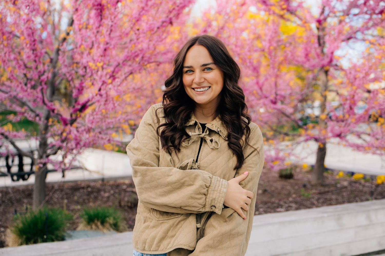 Smiling woman in tan jacket poses with arms crossed before vibrant pink flowering trees.