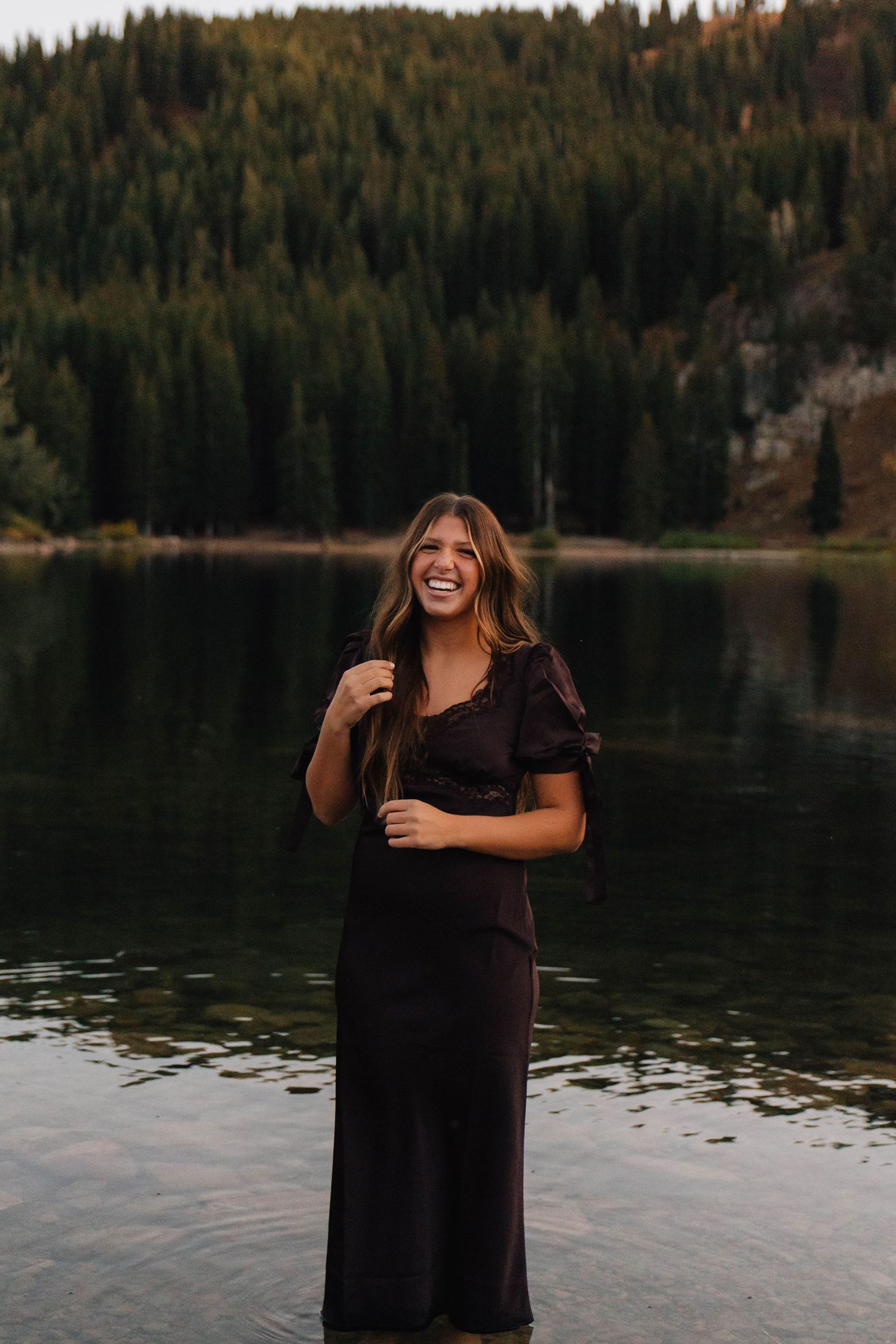 Young woman laughing joyfully while standing in a mountain lake wearing a black midi dress with forest backdrop.