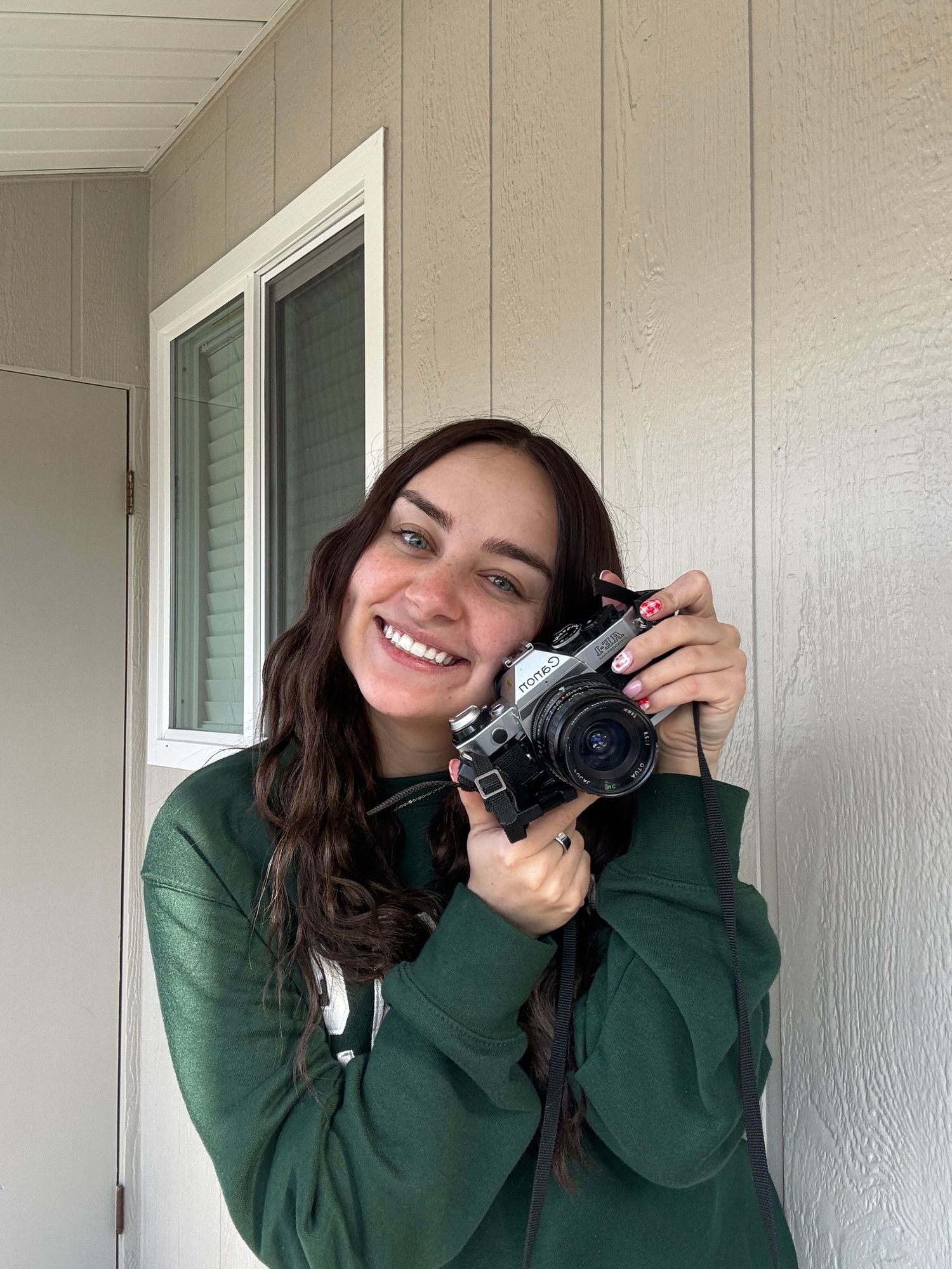 Smiling young woman holding a vintage film camera, wearing a green hoodie, standing outdoors near a building.