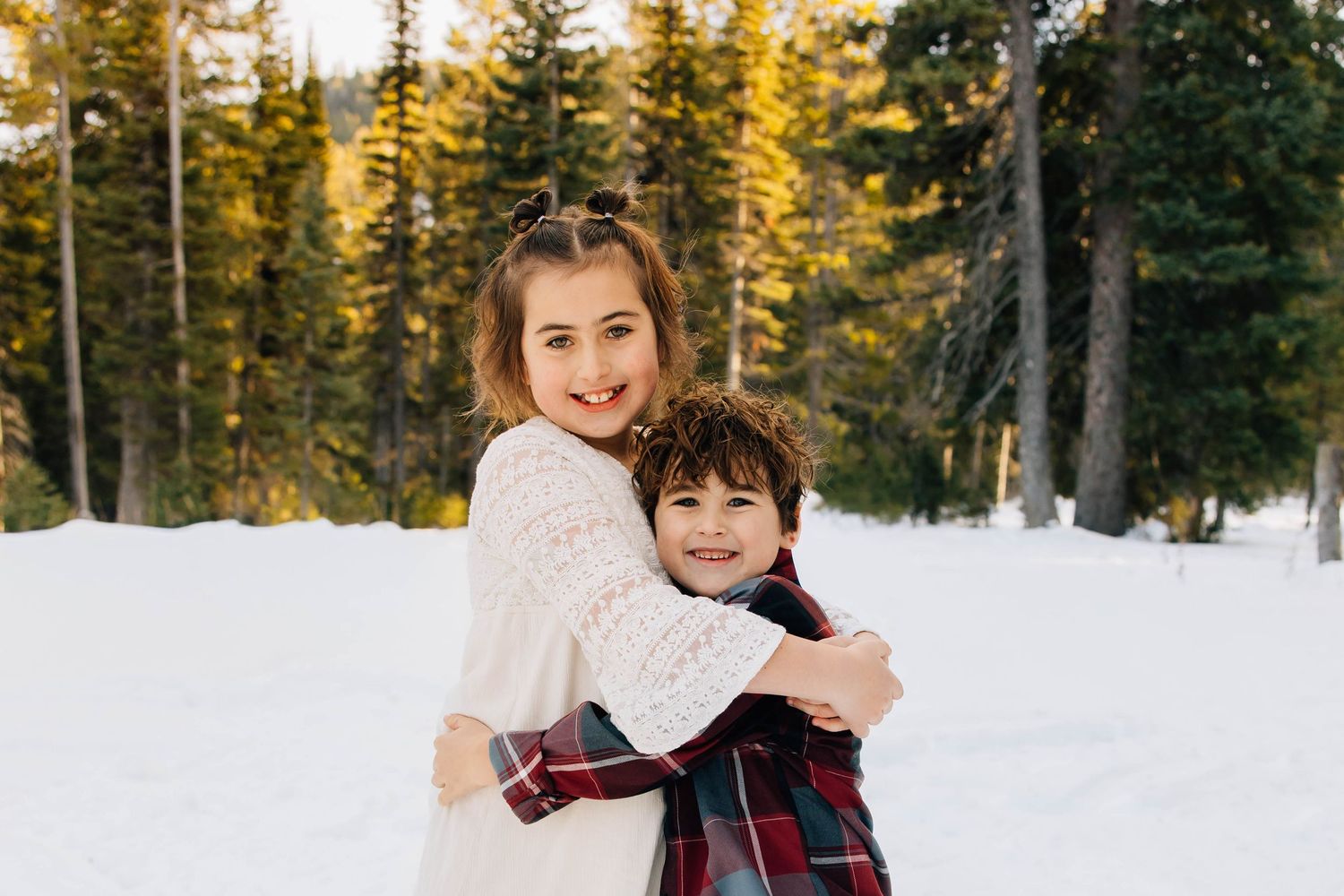 Young girl hugs smiling boy in a snowy forest setting with tall pine trees behind them.