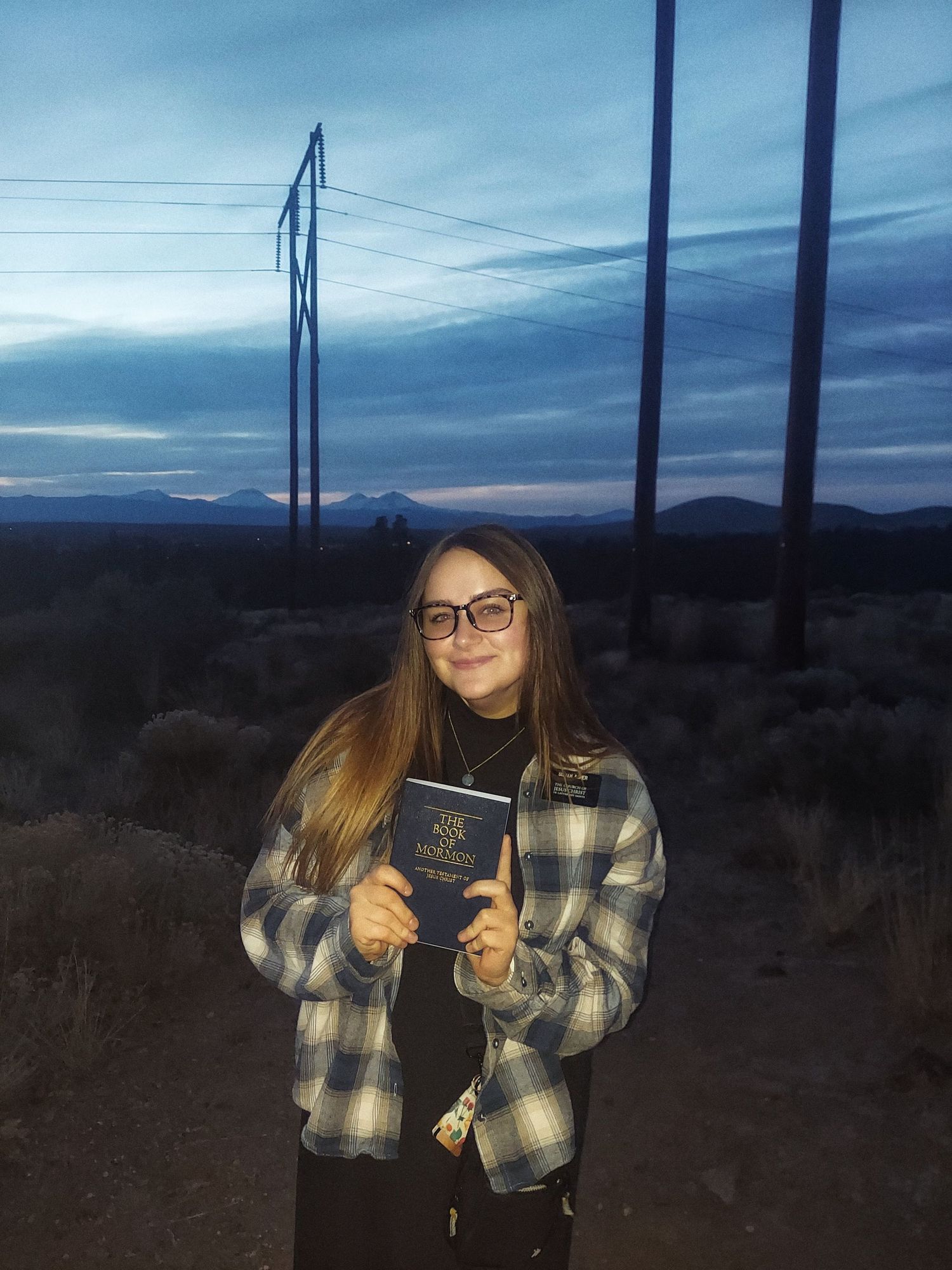 Young woman in plaid jacket holding a book outdoors at dusk with mountains and power lines in background.