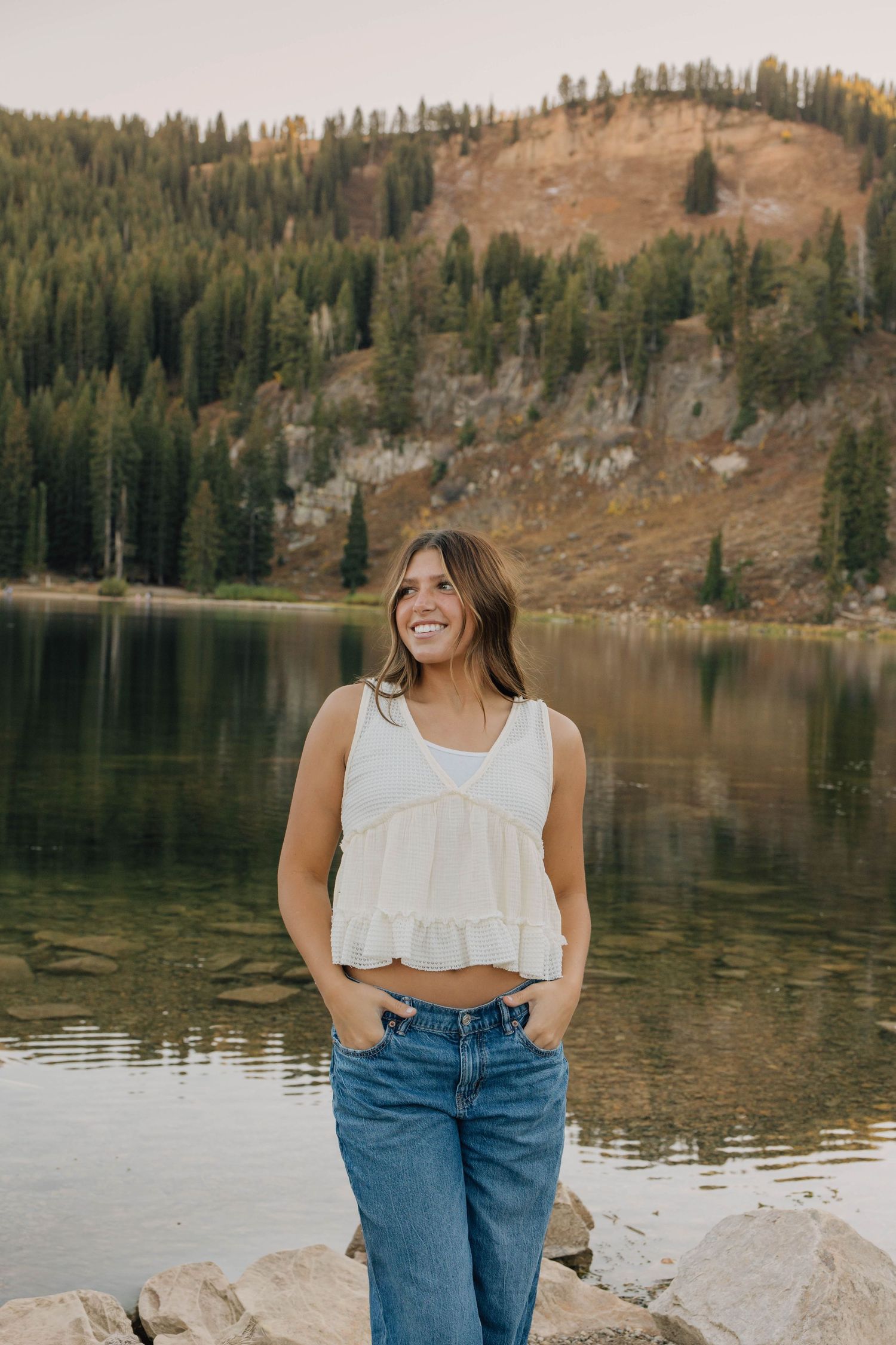 Smiling young woman in white top and jeans stands before a serene mountain lake scene.