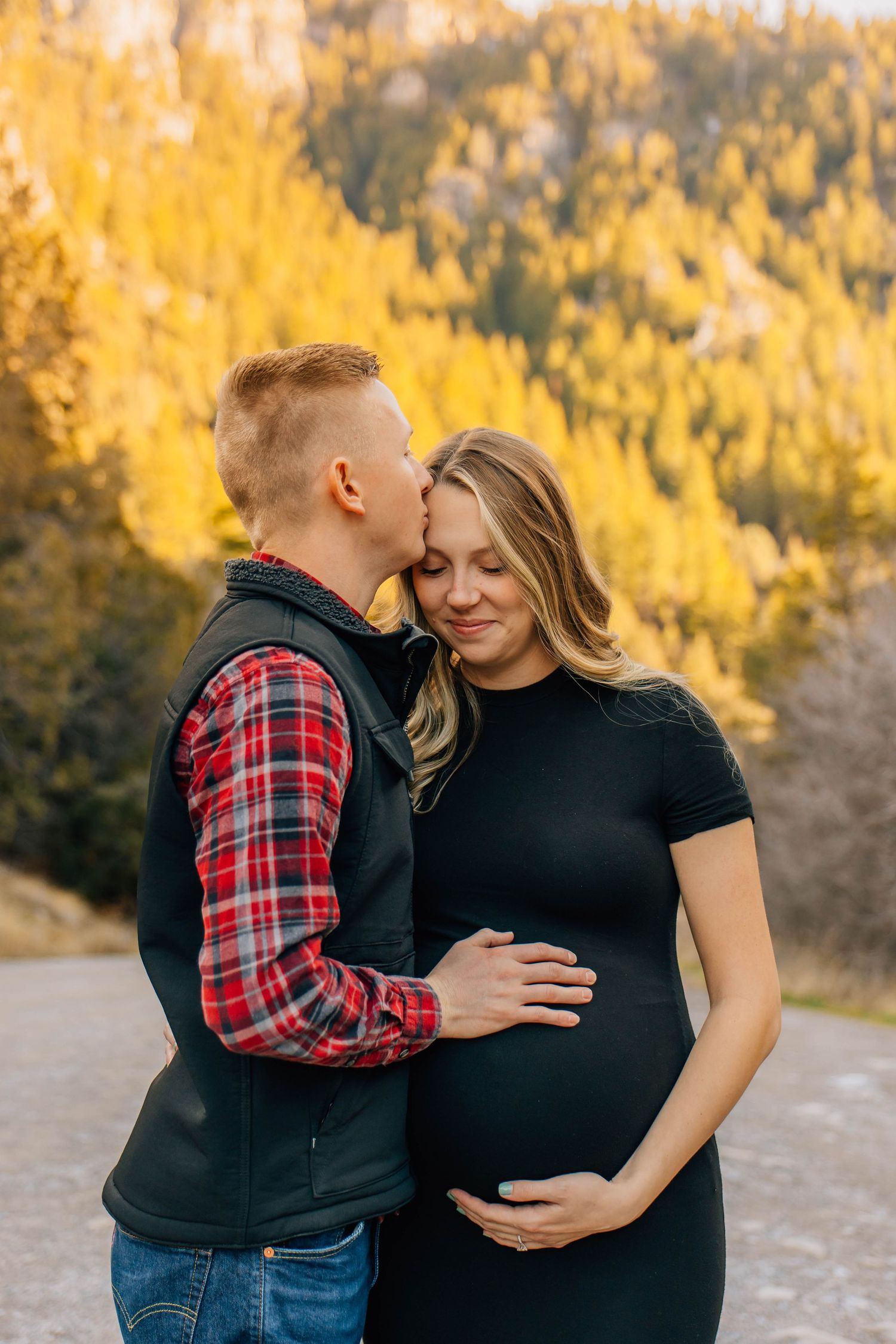 Expecting couple sharing tender moment outdoors in autumn, man kissing womans temple while cradling her baby bump near golden trees.