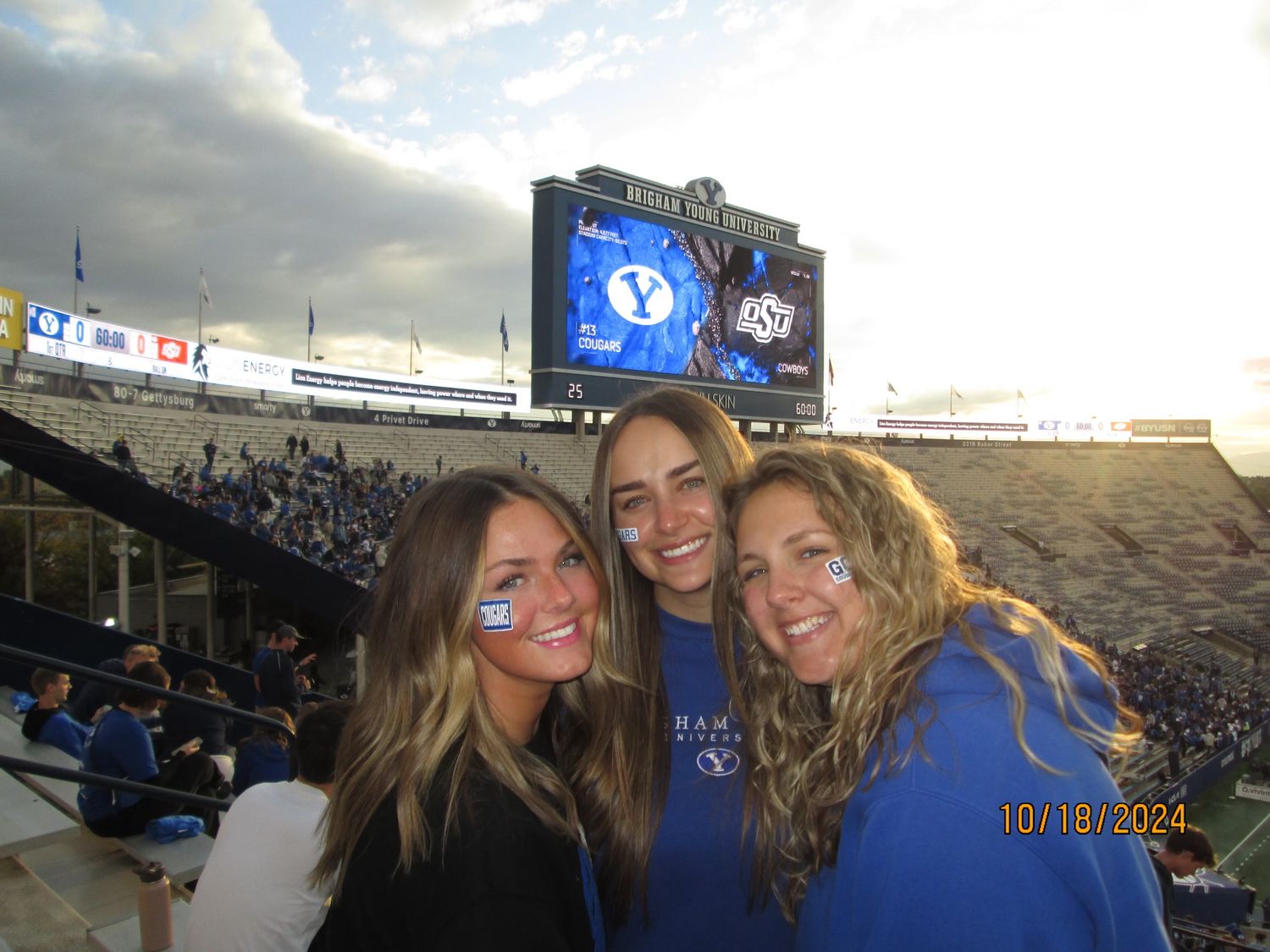 Three female BYU fans smile at a football stadium, wearing blue gear with face stickers, scoreboard visible behind them.