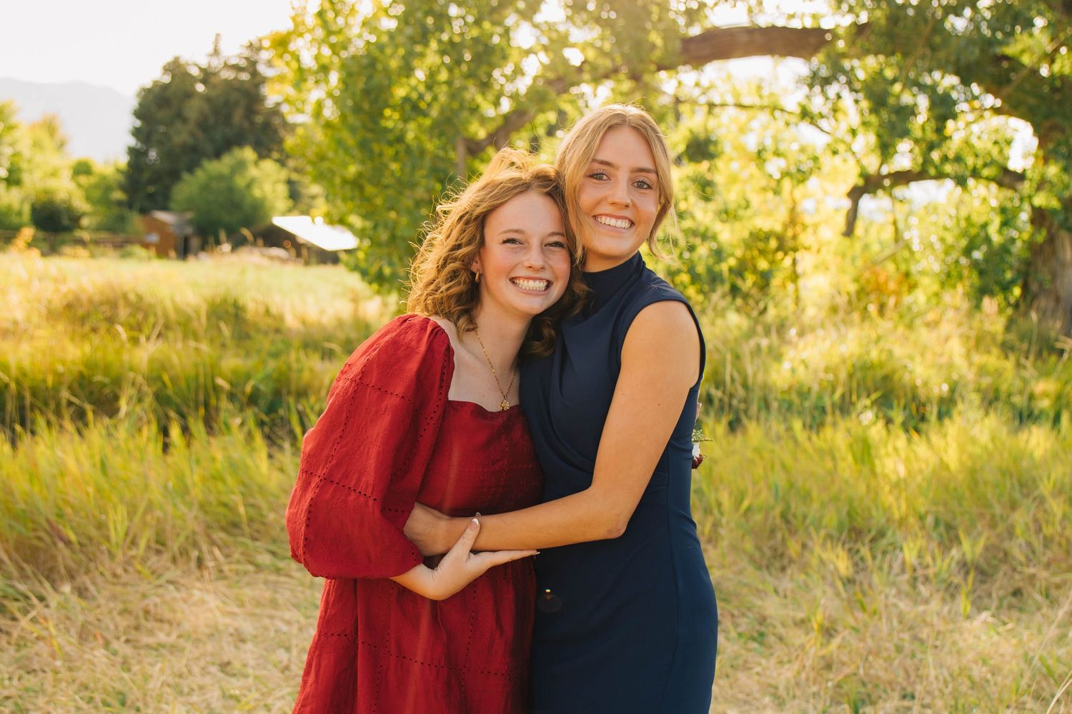 Two smiling women hugging outdoors in sunlit field, one in red dress and one in navy blue dress.