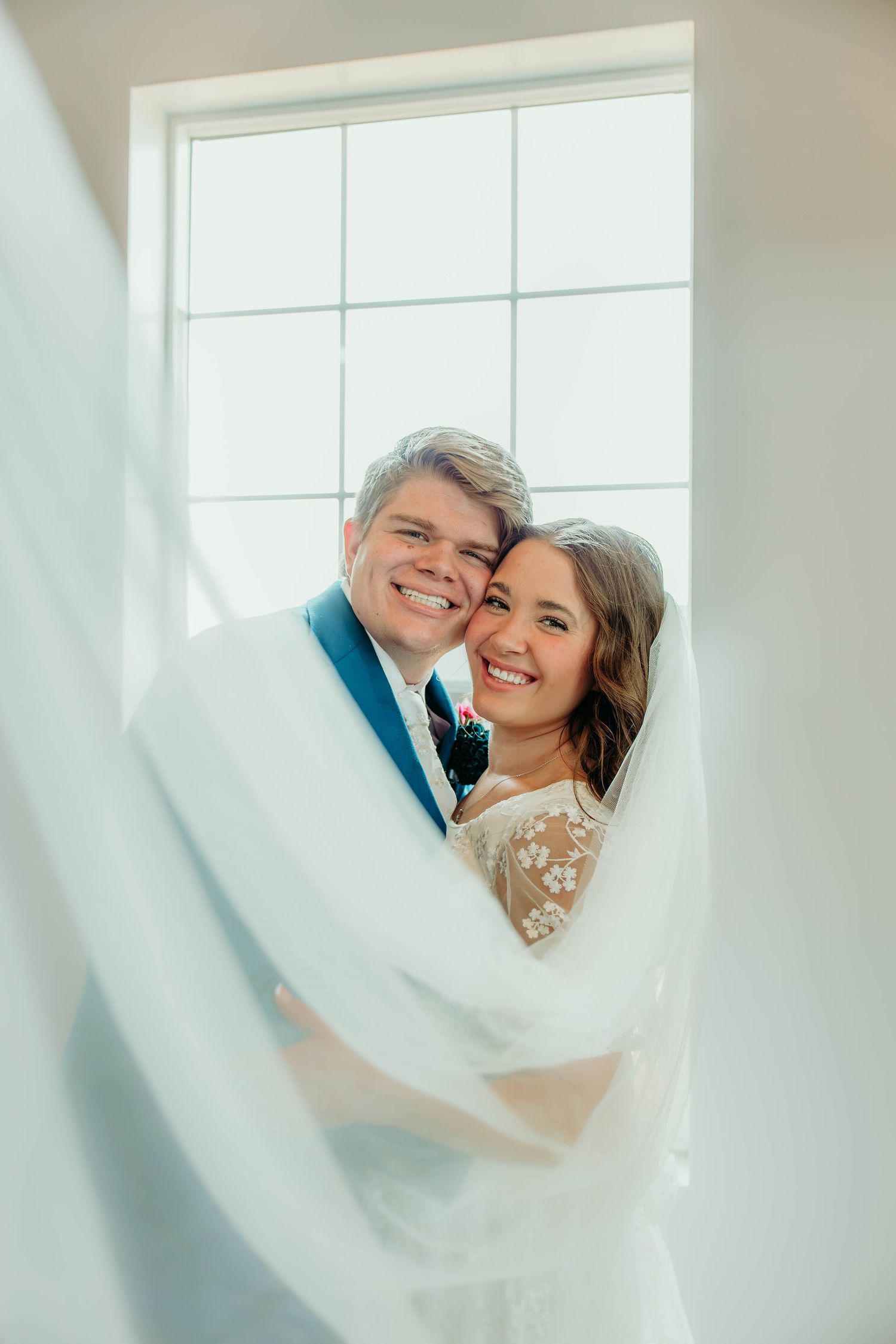 Joyful bride and groom embracing near bright window, bride in lace gown with flowing veil, groom in blue suit.