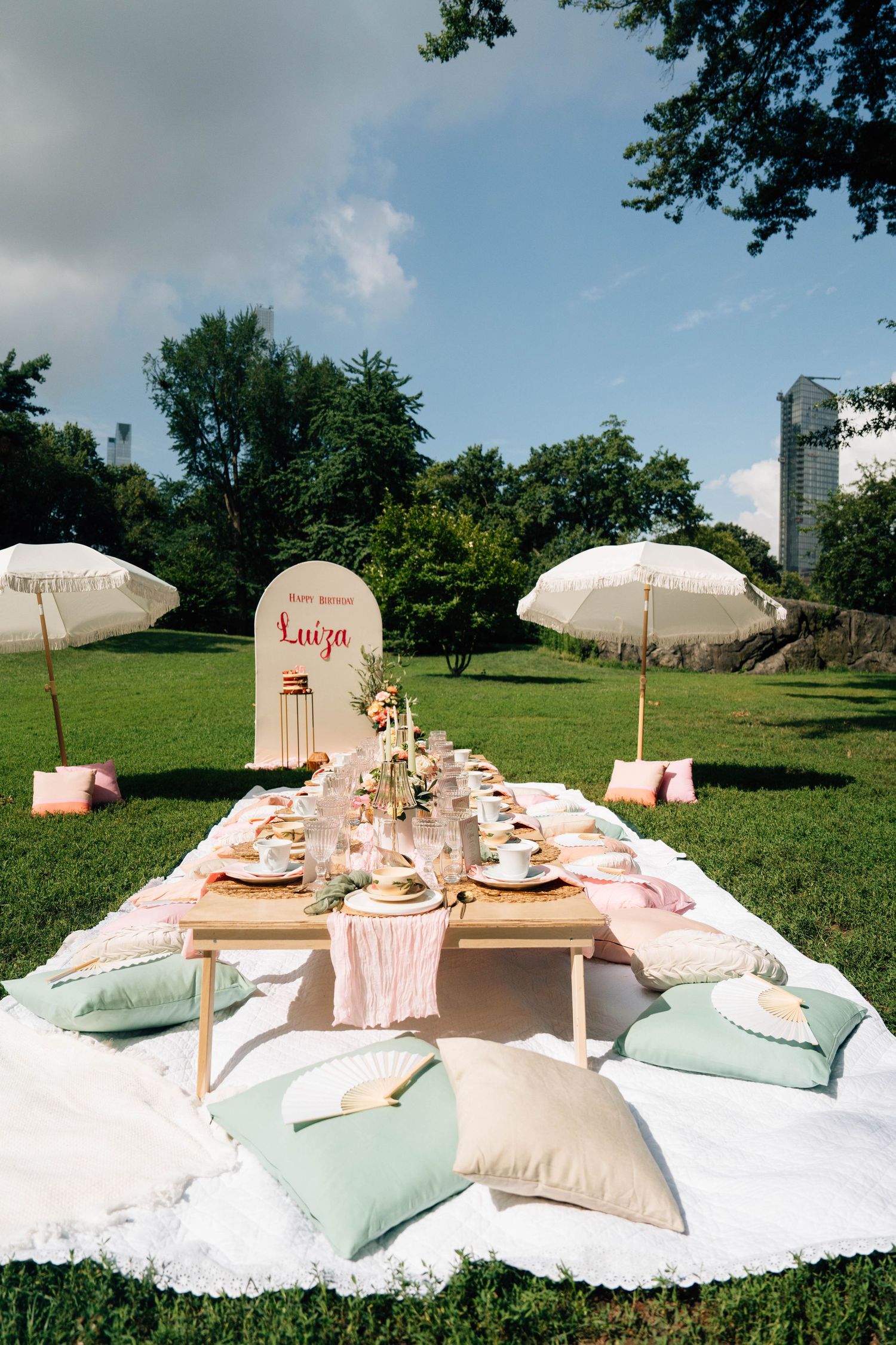 Elegant outdoor picnic setup with white umbrellas, pink and mint cushions, and a long table decorated for a special occasion.