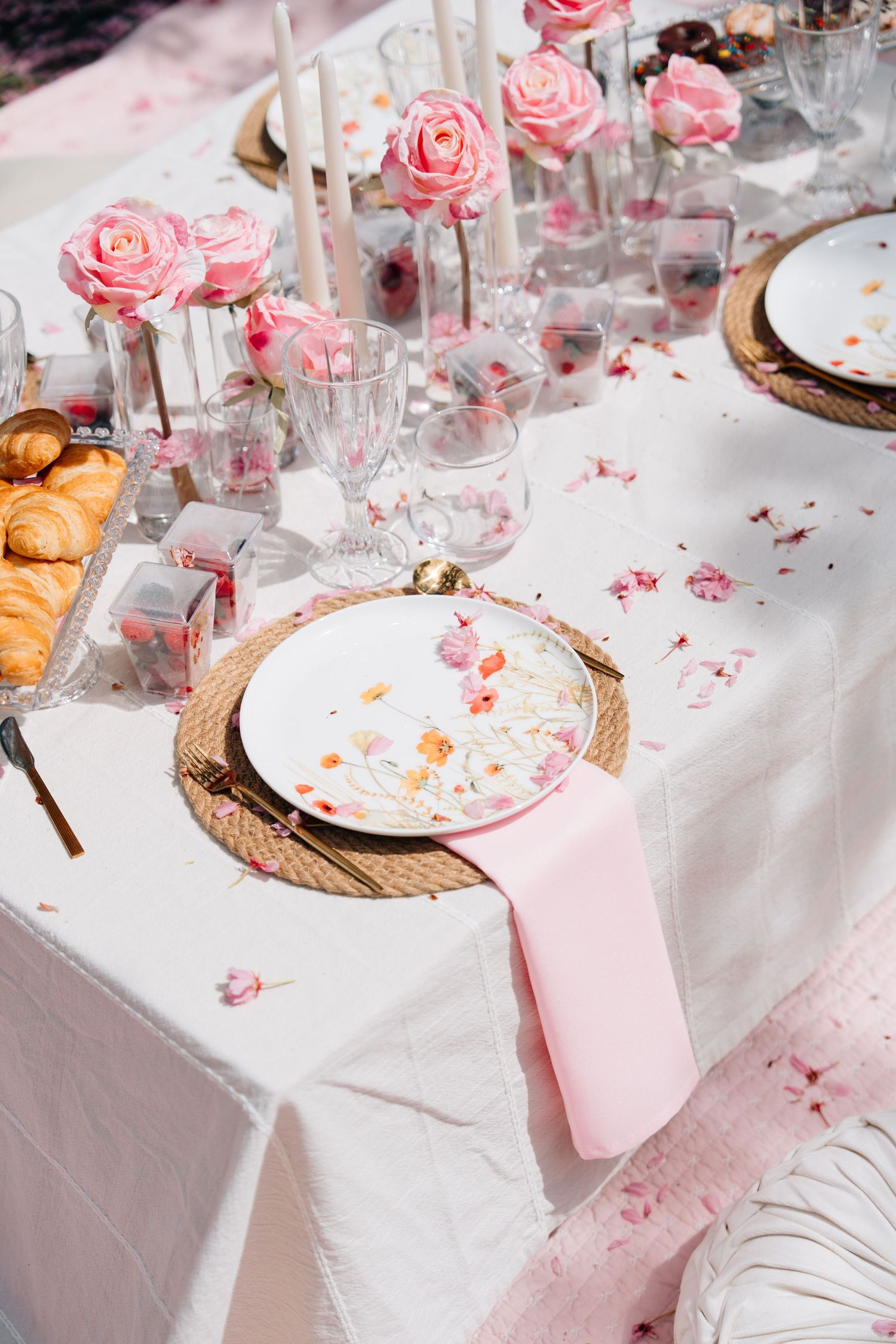 Elegant pink wedding table setting with floral plates, rose centerpieces, candles, and croissants on white linen.