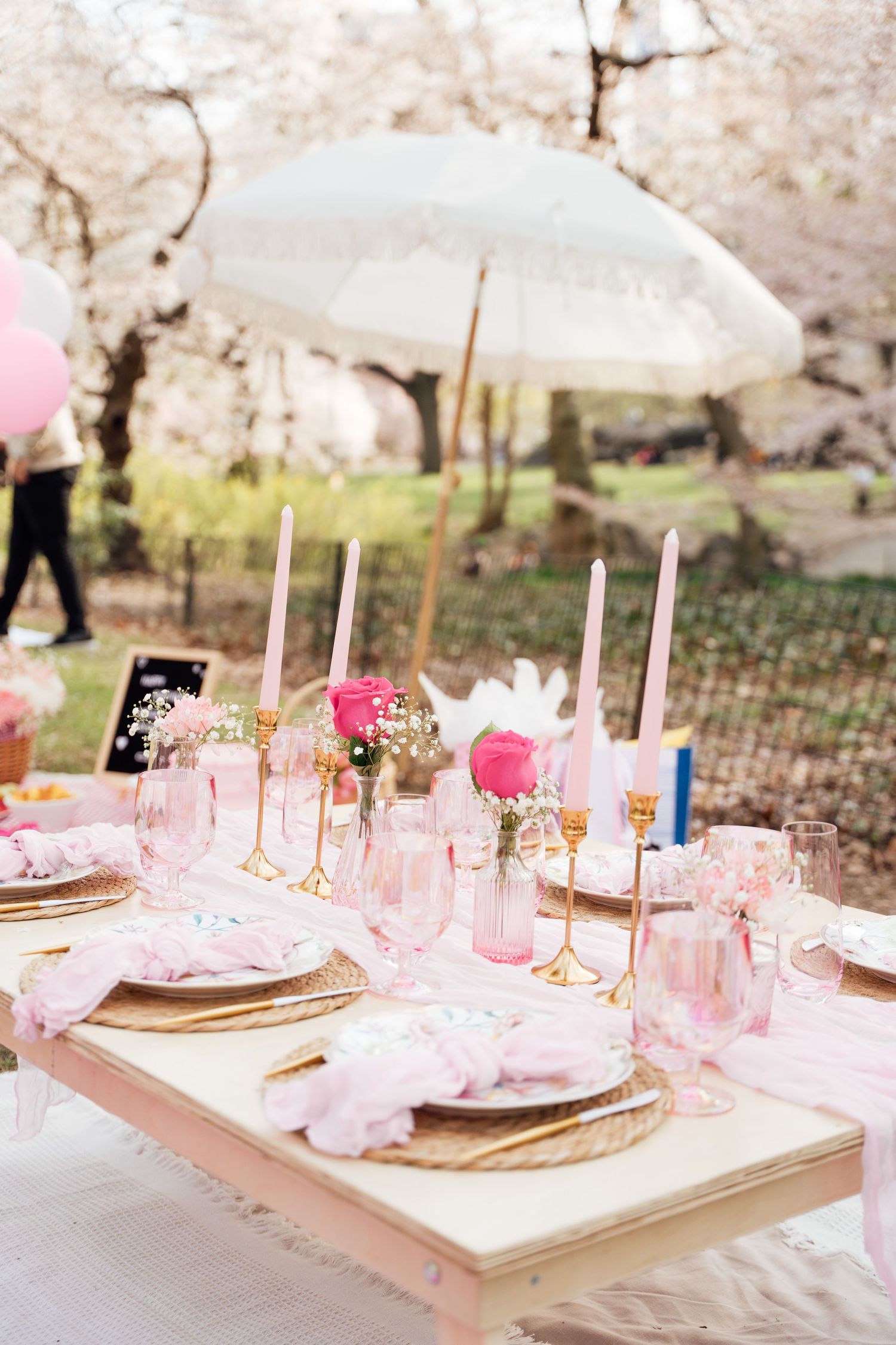Pink cherry blossom picnic table setting with gold candlesticks, pink tapers, floral arrangements, and white umbrella outdoors.