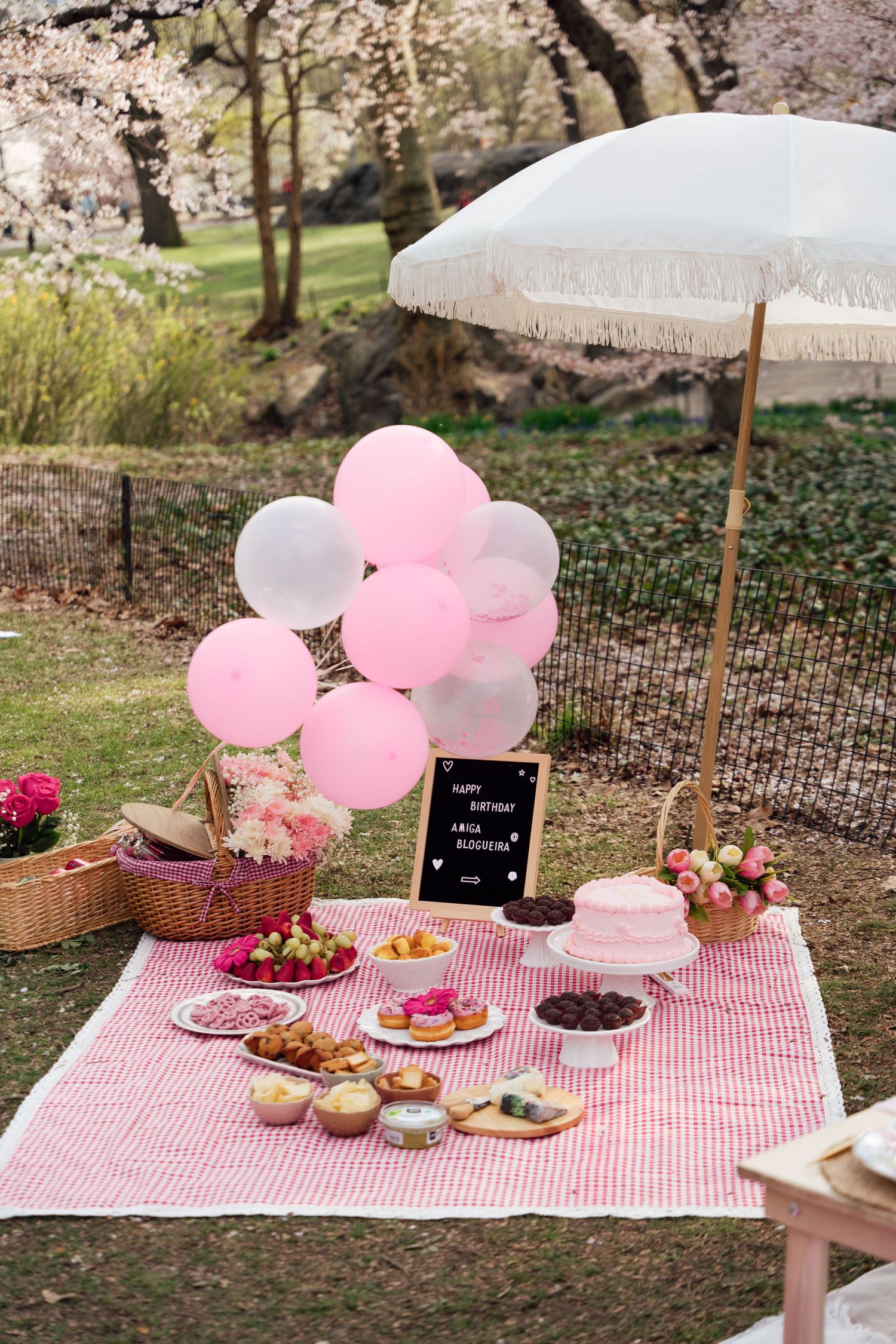 A picturesque outdoor picnic setup with pink and white balloons, checkered blanket, and treats under cherry blossom trees.