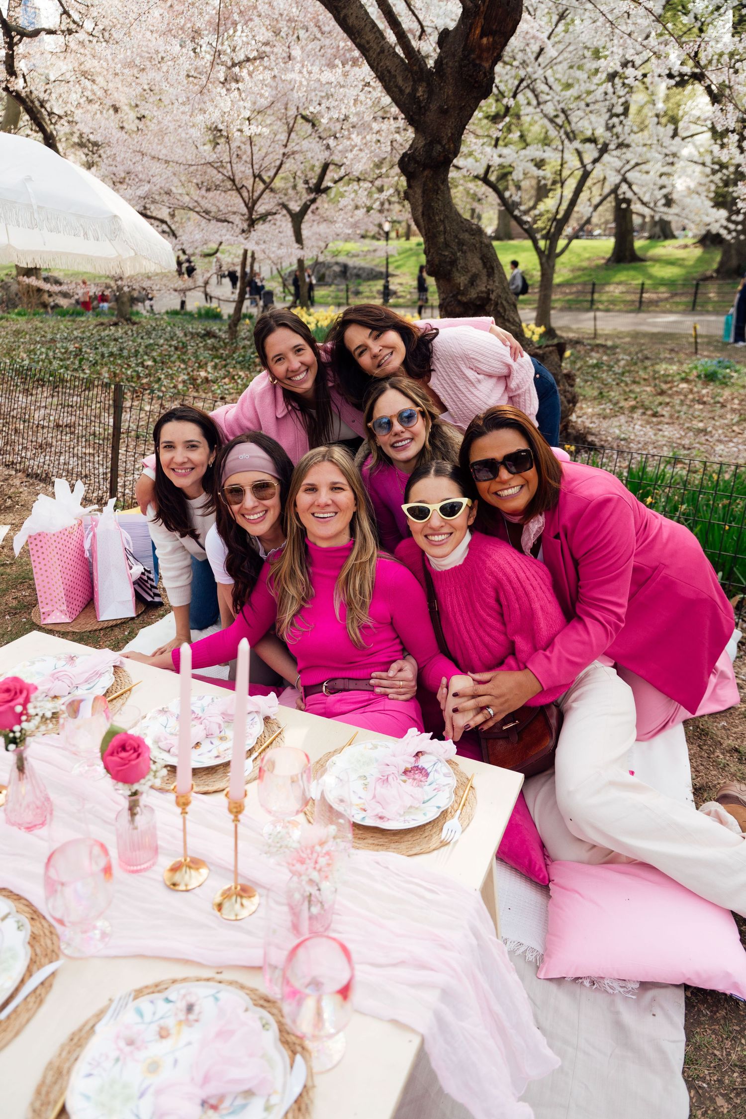 A spring picnic gathering under cherry blossom trees with guests wearing pink outfits enjoying food on a decorated table.
