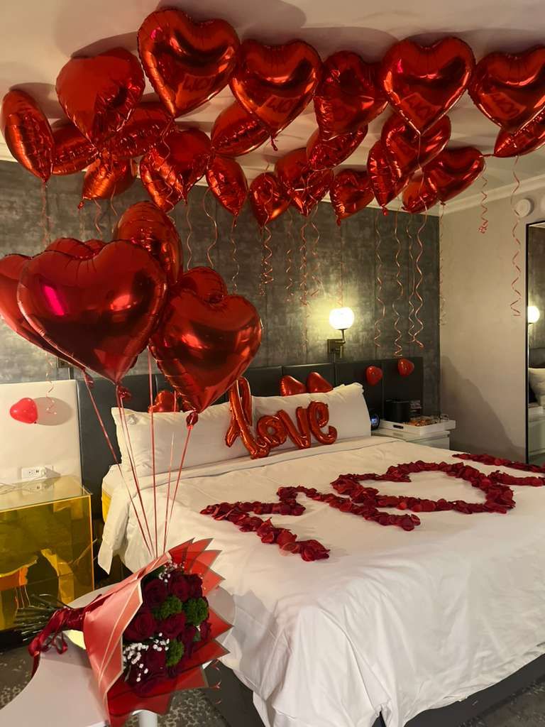 A romantic hotel room decorated with red heart balloons and rose petals scattered on a white bed for Valentine's Day.