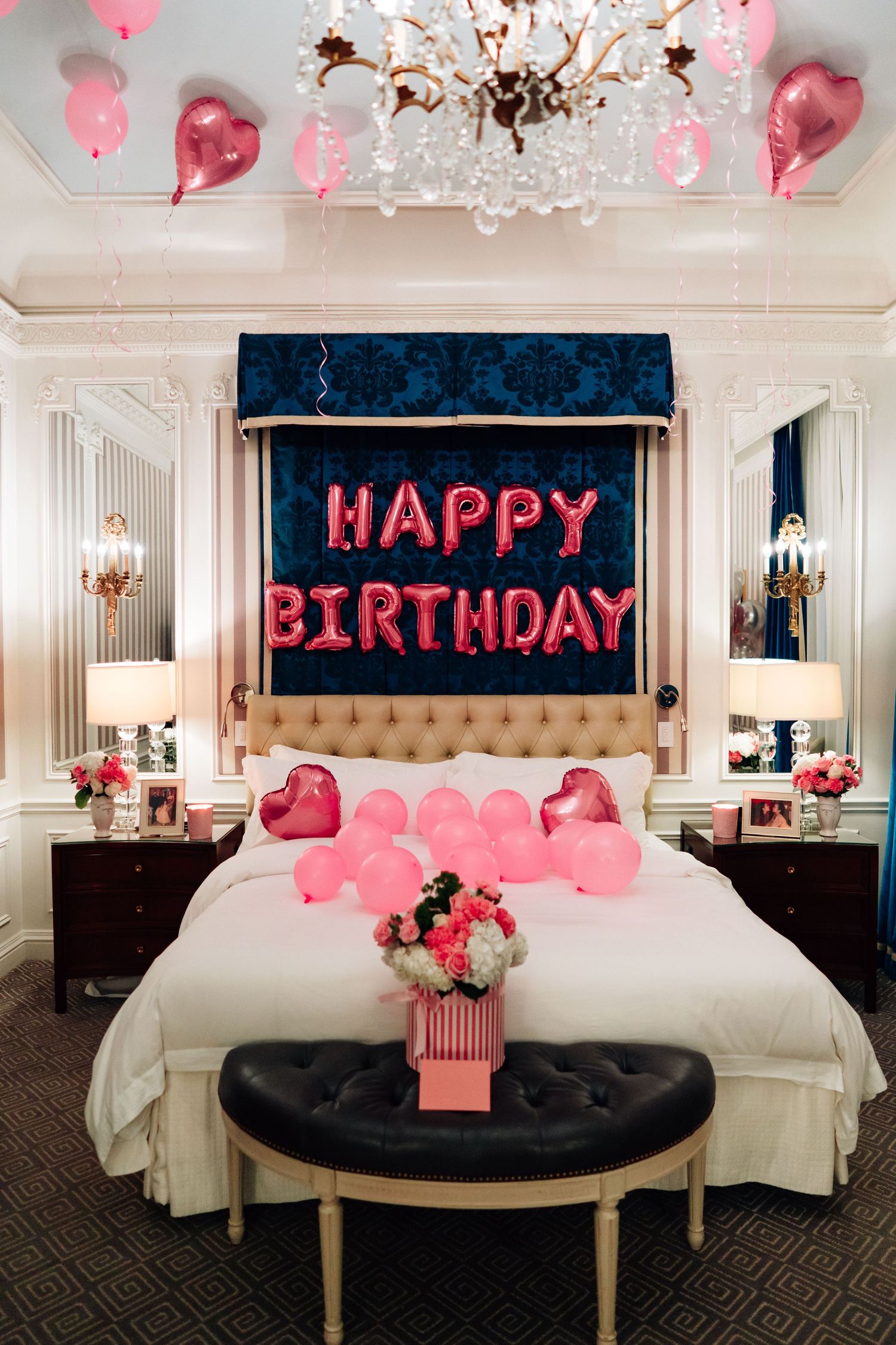 Romantic hotel room decorated with pink heart balloons, white bedding, and 'Happy Birthday' text display for celebration.