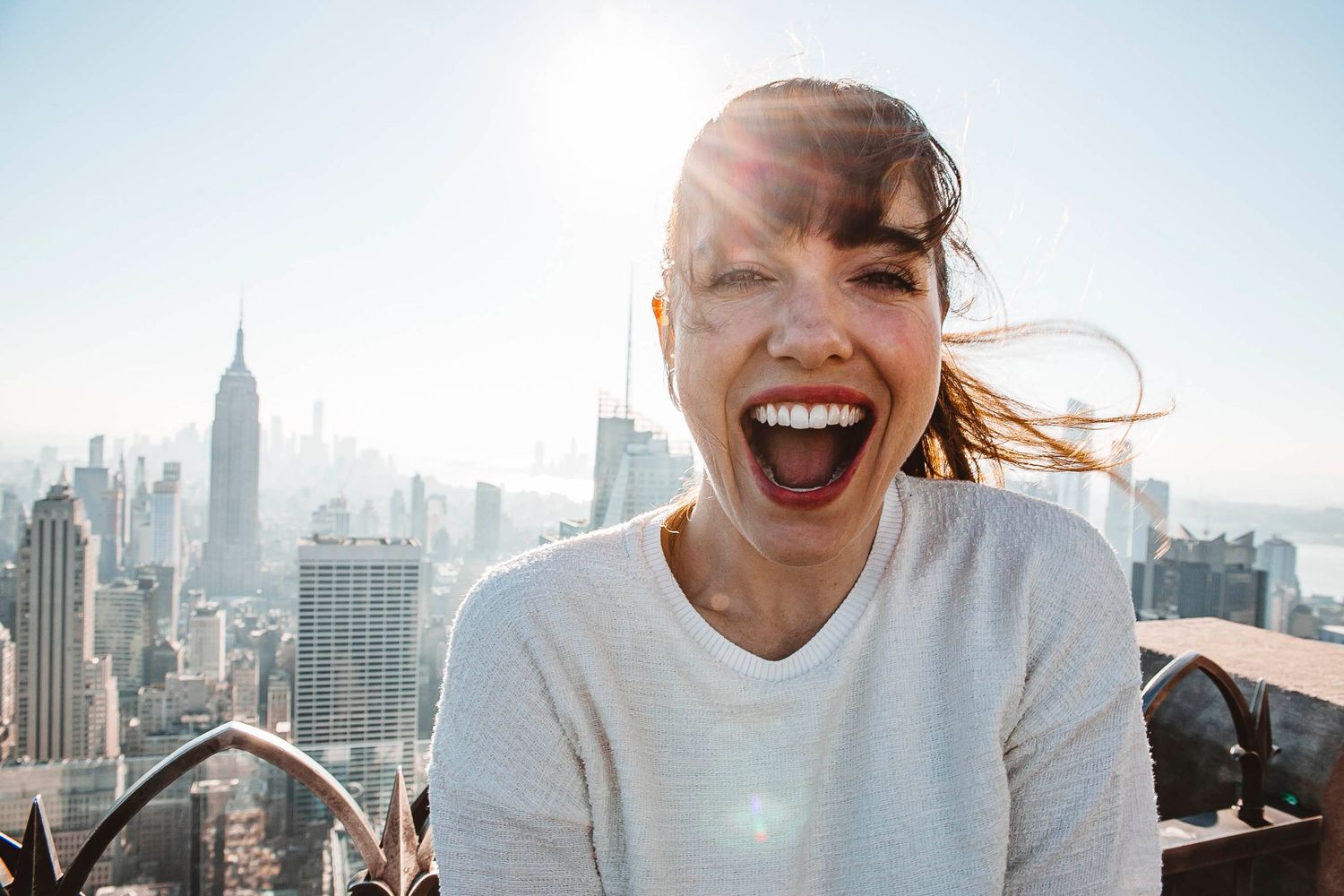 A joyful person laughs while enjoying a rooftop view of the New York City skyline with the Empire State Building visible.
