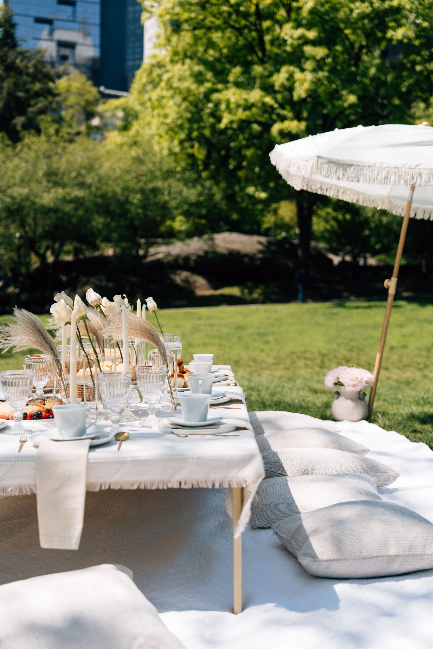Elegant outdoor boho picnic setup with low table, floor cushions, pampas grass, glassware, and fringed umbrella on green lawn.