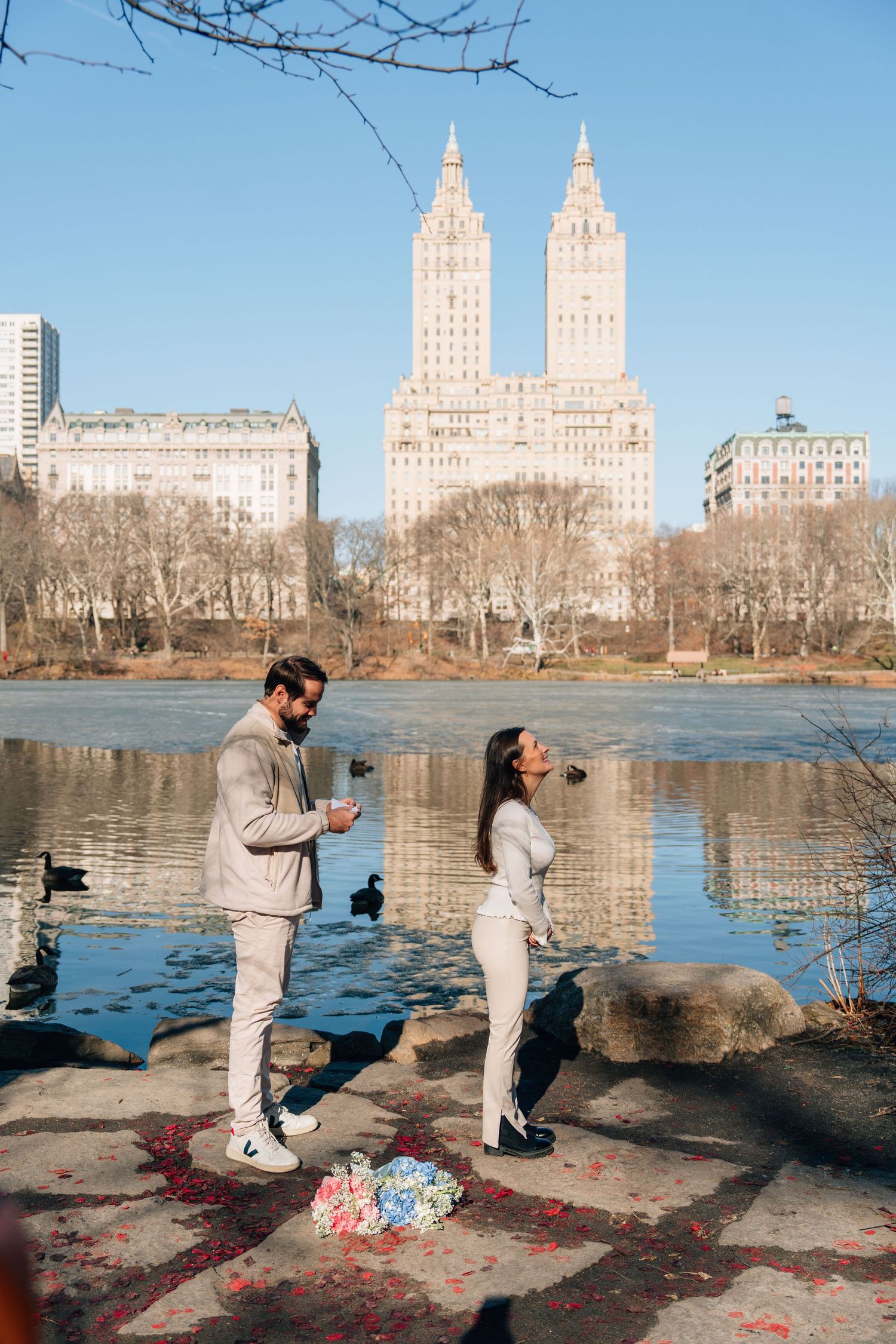 Couple sharing a romantic proposal moment by a lake in Central Park, NYC, with iconic San Remo building in background.