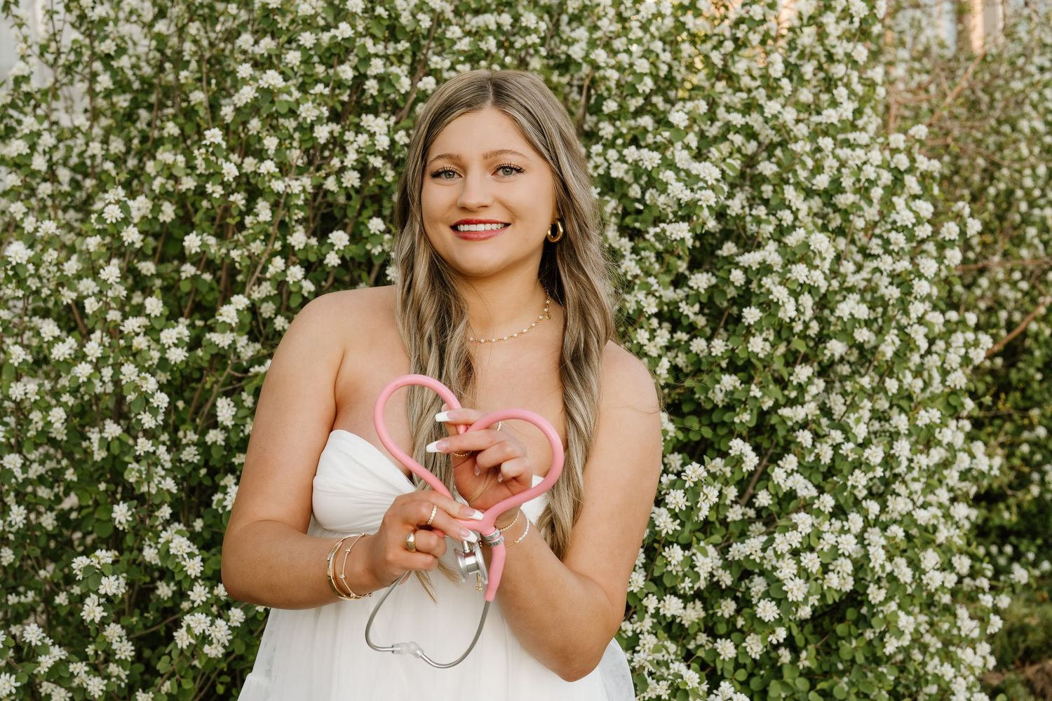 A University of Utah graduate in front of a flowering bush and her stethoscope in the shape of a heart; Graduate poses