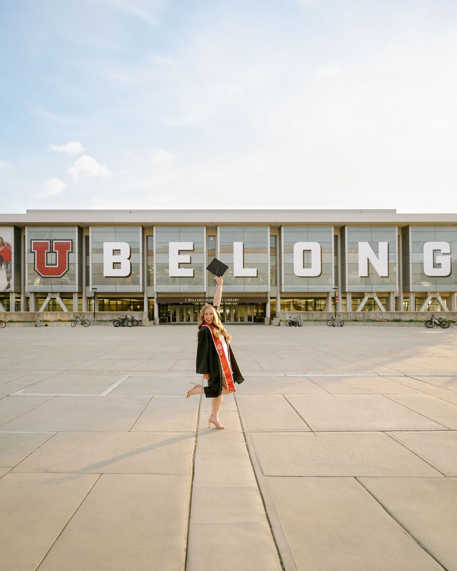 A University of Utah graduate raising her cap in her gown in front of the library