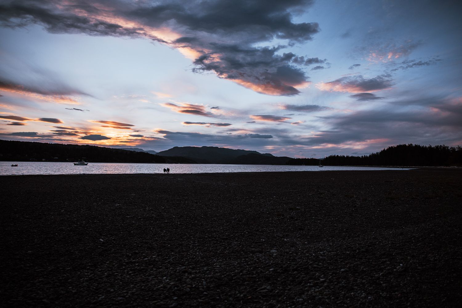 Dramatic sunset over a lake with dark silhouetted mountains and moody clouds reflecting pink and orange hues in the sky.