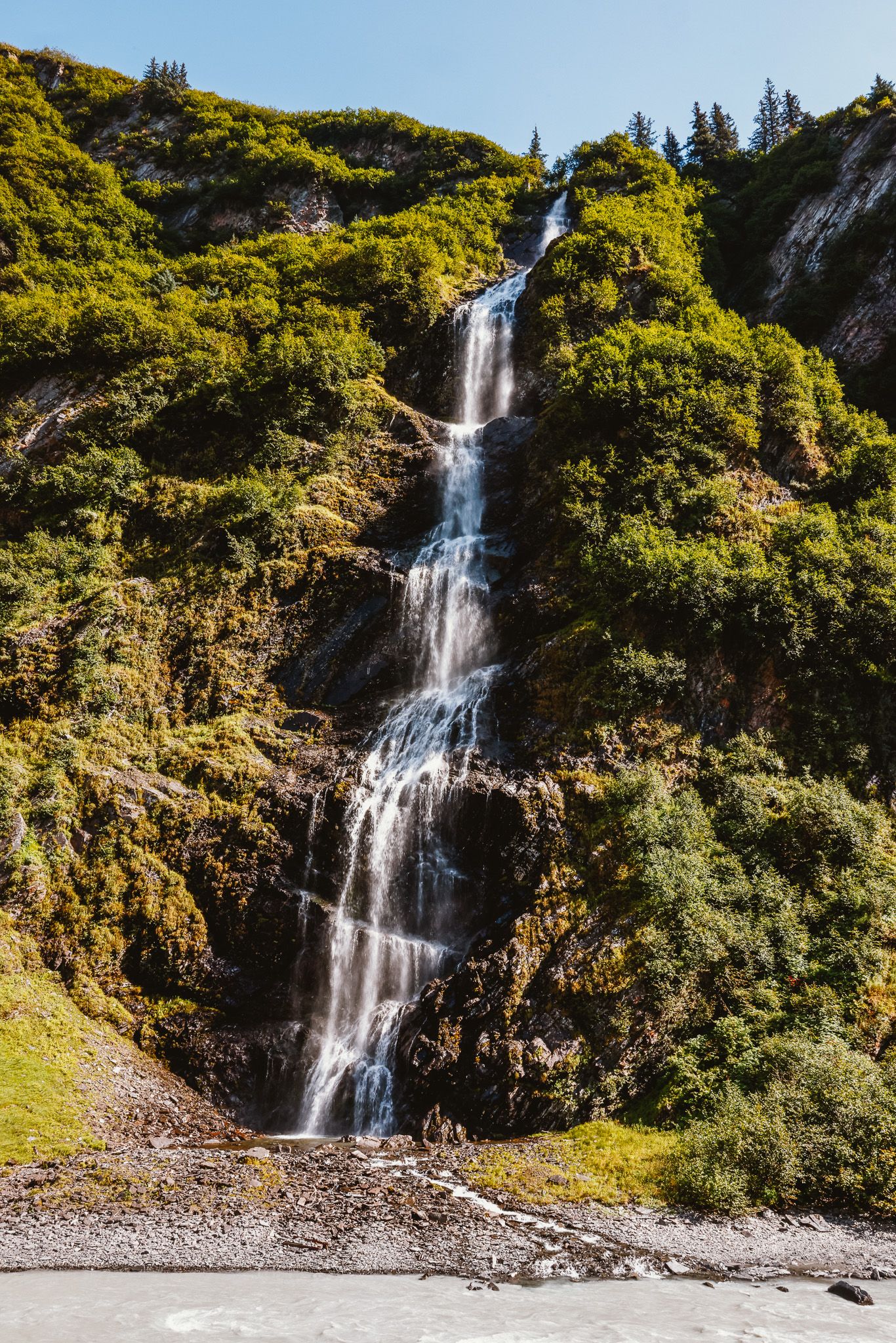 Tall cascading waterfall flows through green alpine landscape on sunny mountainside.