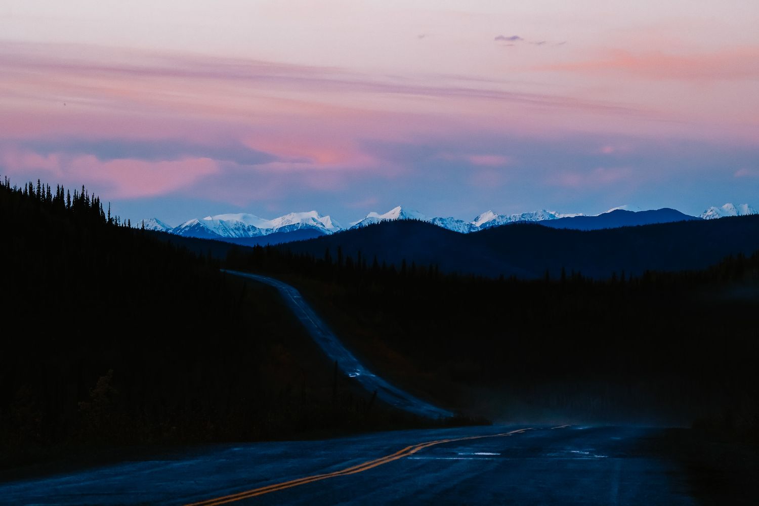 Winding mountain road at dusk with snow-capped peaks and pink-purple sky in the background.
