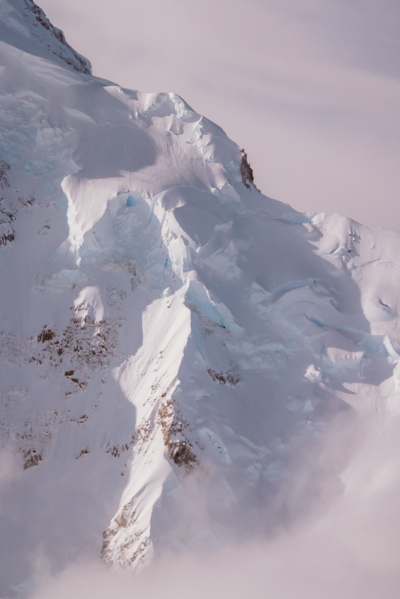 Aerial view of snow-covered mountain peaks with exposed rocky outcrops under cloudy winter skies.