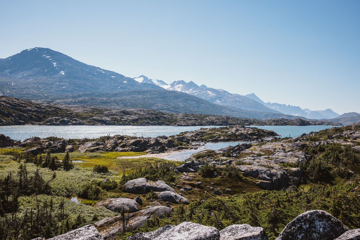 Rocky coastal landscape with snow-capped mountains and calm waters under clear blue skies.