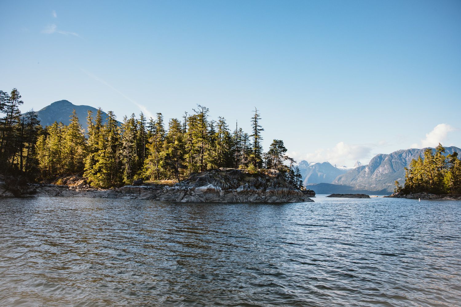 Small rocky island with evergreen trees stands in a mountain lake surrounded by rugged peaks on a sunny day.