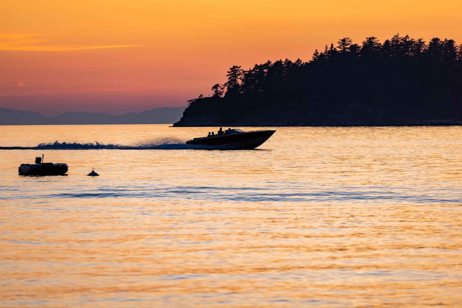 Silhouettes of boats against a vibrant orange sunset over calm waters with a forested island in the background.
